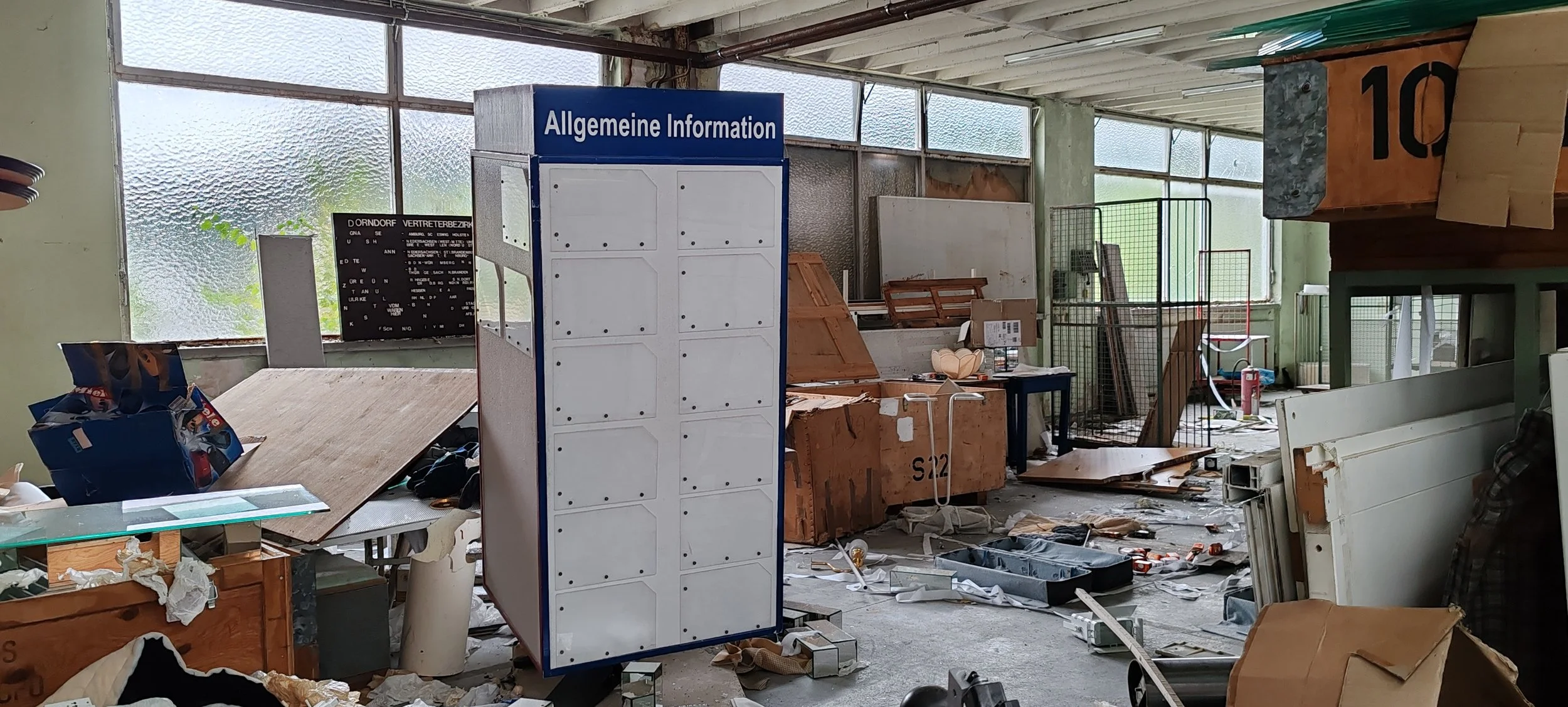 Unaufgeräumter Raum mit Müll, Holzstücken, leeren Boxen, einer Info-Tafel, einem Feuerlöscher und zerbrochenen Gegenständen. Fenstern mit verdrecktem Glas im Hintergrund.
