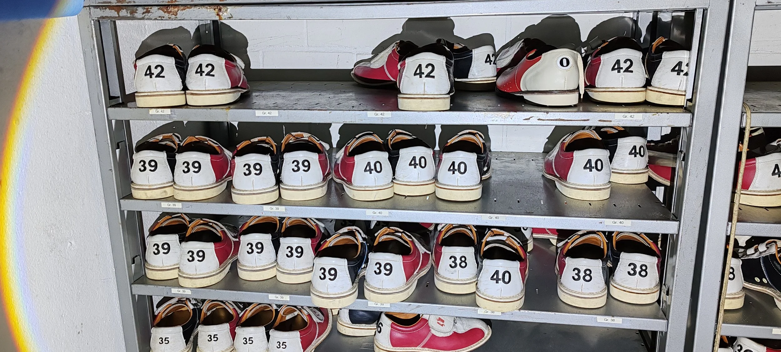 Mehrstöckiges Schuhregal mit weißen Kinderschuhen in verschiedenen Größen, nummeriert von 35 bis 42, in einer Turnhalle oder Sporthalle.