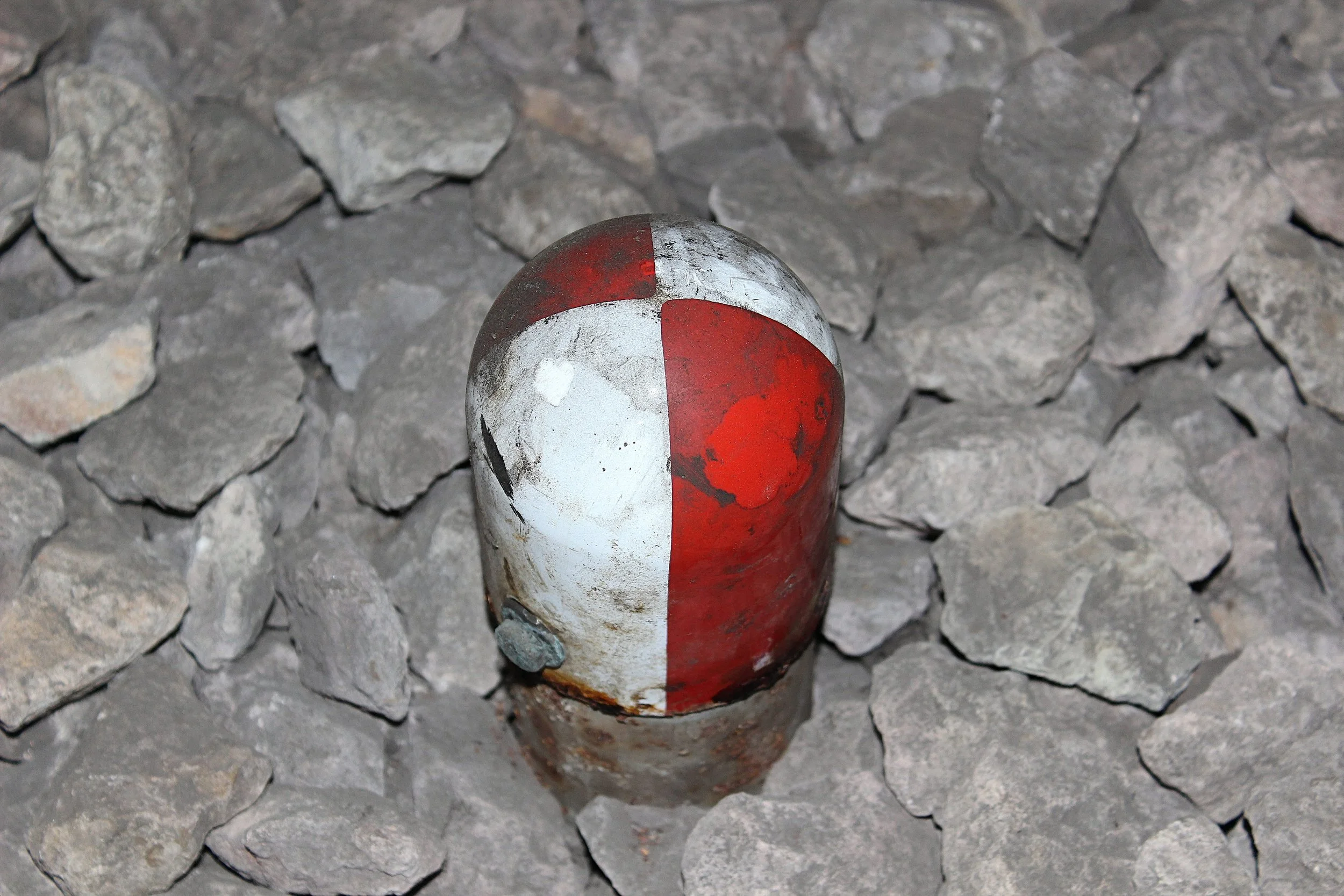 Ein verrosteter, halbweiß, halbroter Betonschweller auf einem umgebenden Graustoff aus Steinen.