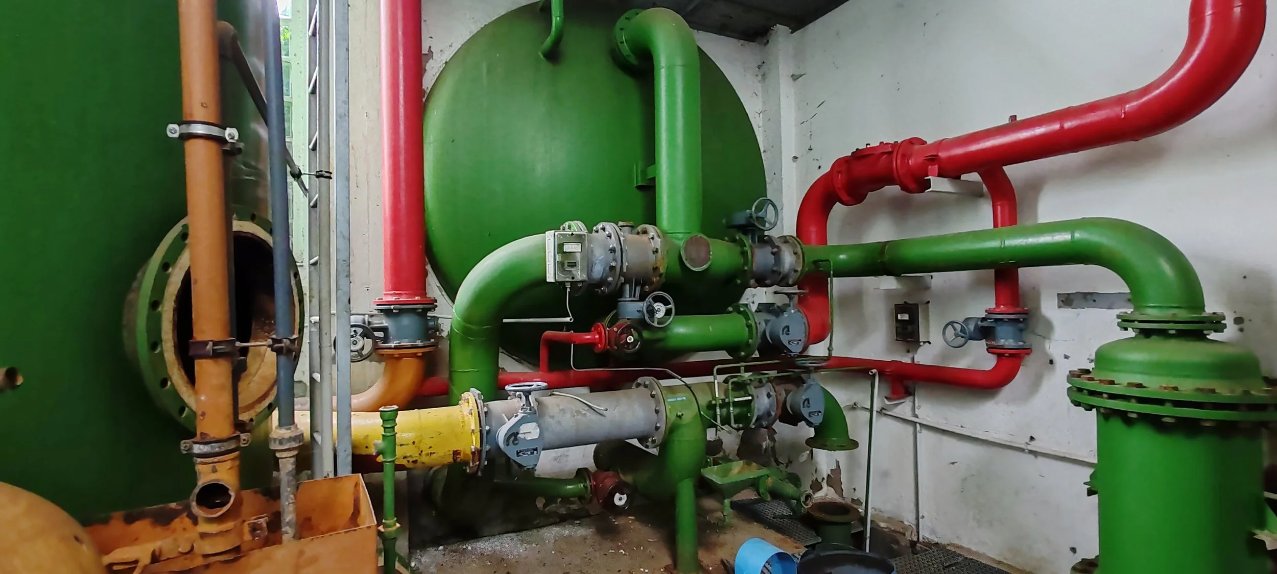 Installierte Wasserleitungen und Pumpen in einem Technikraum, mit roten, grünen und gelben Rohren, samt Ventilen und Messgeräten.