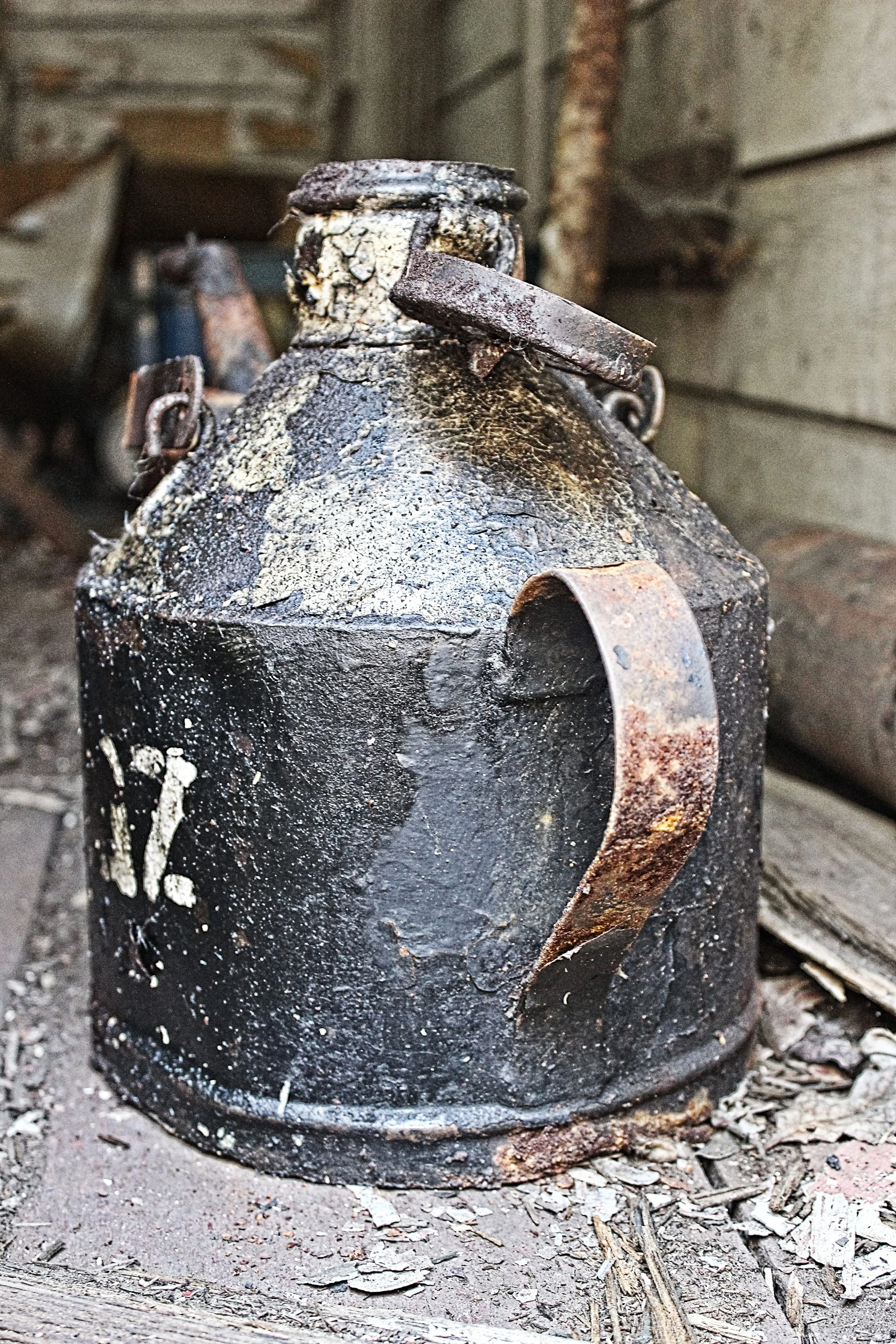 Ein alter, verrosteter Metallkessel mit einer großen Grifföffnung und Metalllasche auf dem Deckel, sitzt auf dem Boden in einer Scheune oder verlassenen Hütte.