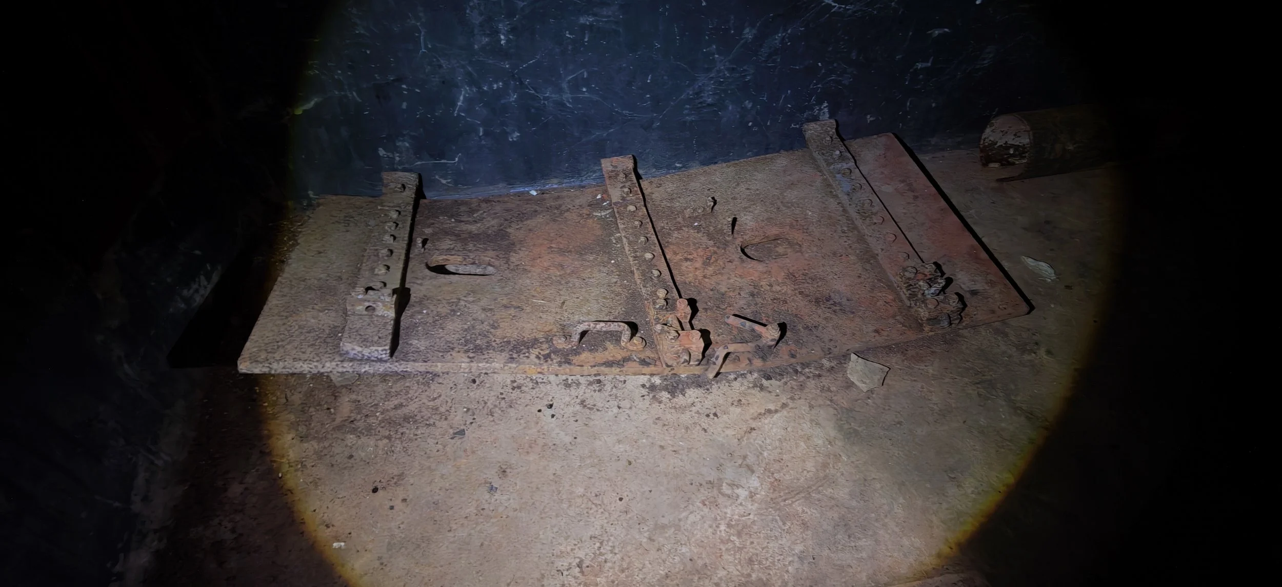 Ein rostiges, beschädigtes Metallstück liegt auf einem Boden aus Schmutz und Staub, in einer dunklen Umgebung mit einer Lichtquelle, die auf das Metallstück gerichtet ist.