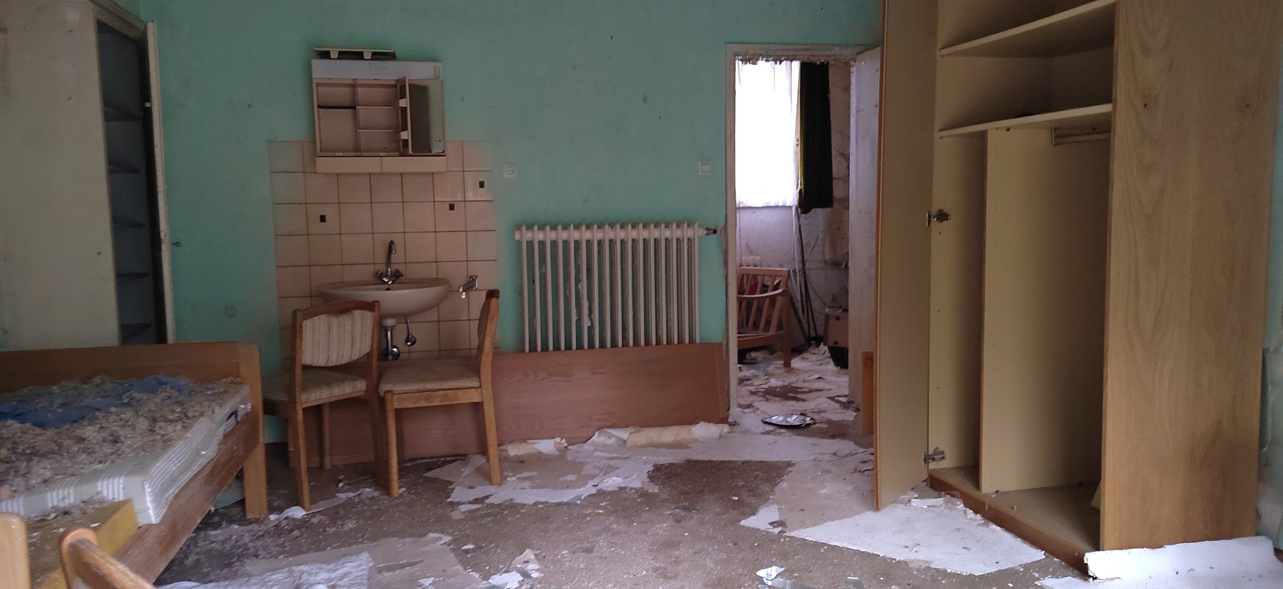 Ein unordentliches, vernachlässigtes Zimmer mit Kisten, Betten, Stühlen, einem Wasserhahn und einer Heizung. Der Boden ist schmutzig und mit Papier bedeckt.