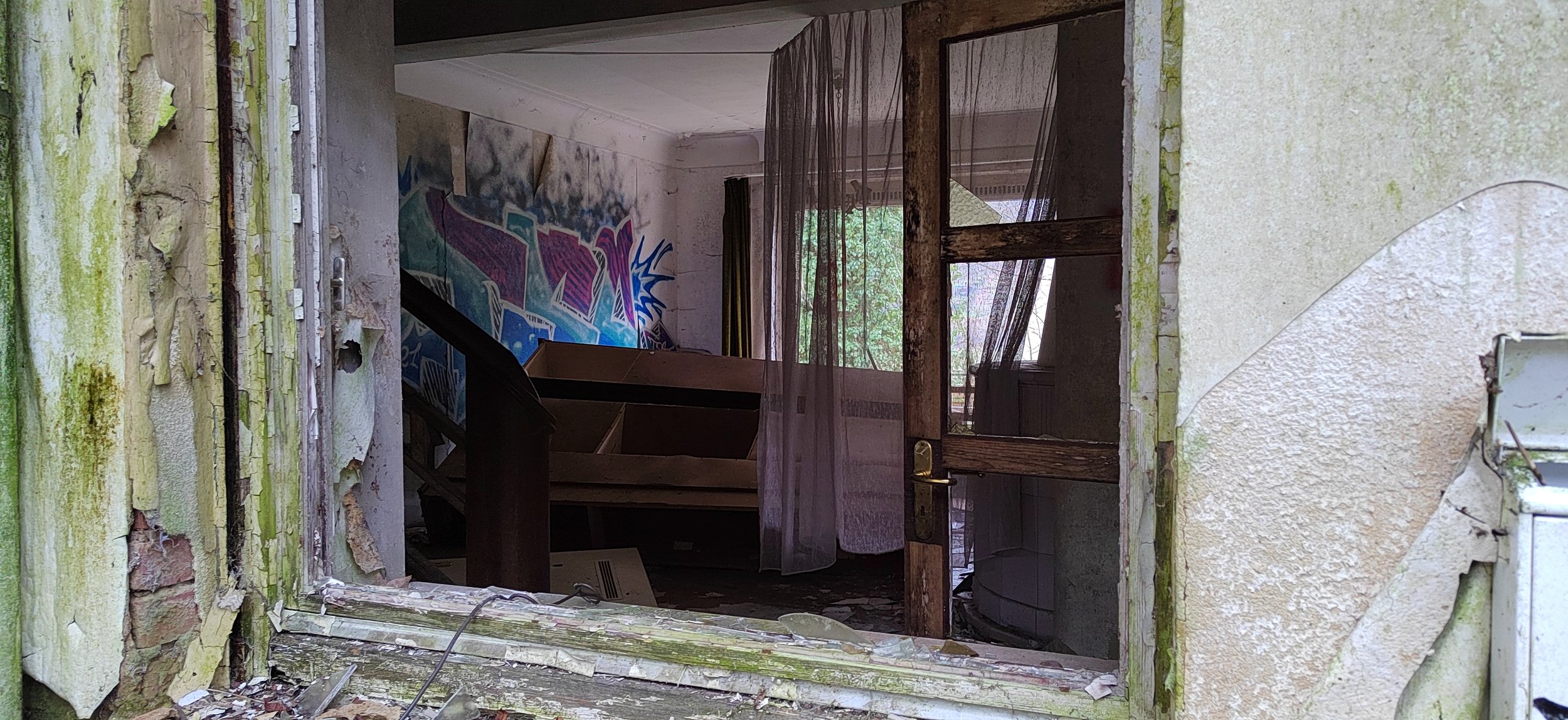Verlassene, verwahrloste Innenraum eines alten Hauses, durch ein zerbrochenes Fenster sichtbar, mit Graffiti an der Wand und vollgestelltem Raum.