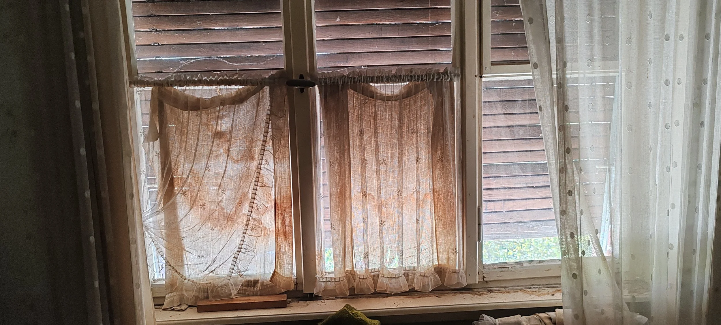 Blick durch einen Fensterrahmen auf eine Fensterbank mit halb zugezogenen, beige-braunen Vorhängen und einem Holzboden vor einem Holzzaun draußen.