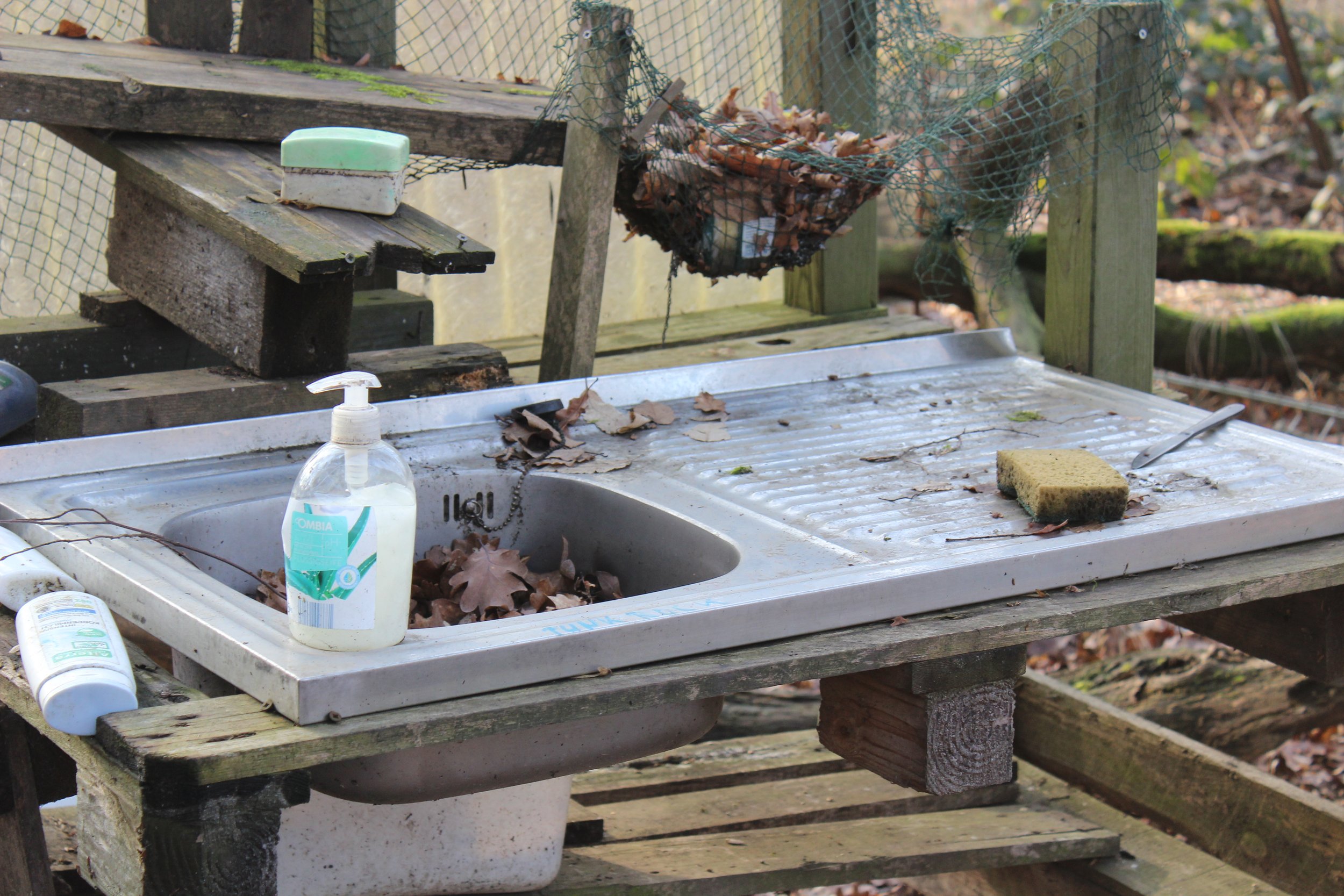 Ein abgestelltes Küchenspüle im Freien mit Laub und Schmutz, daneben eine Seife und ein Schwamm, umgeben von Holzstücken und Gartenmaterialien.