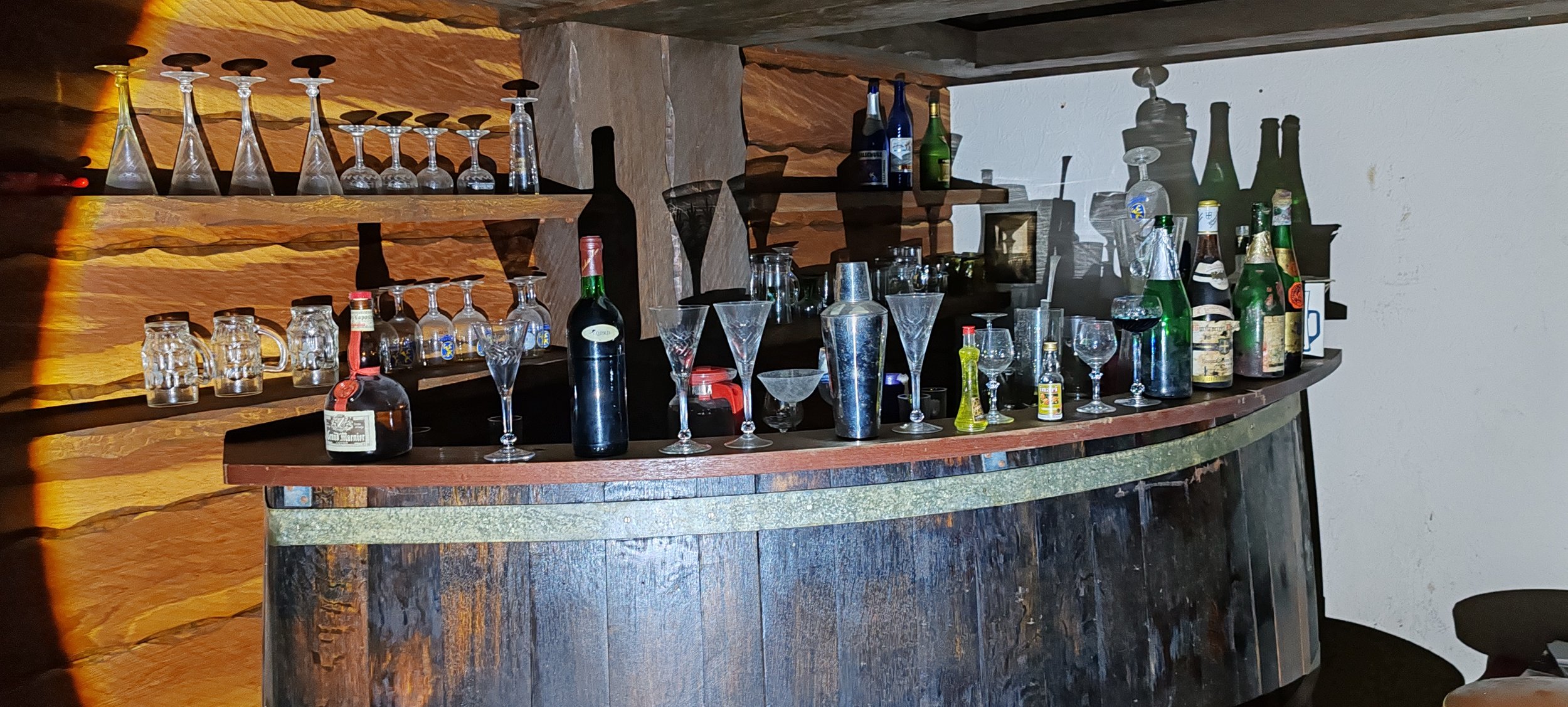Eine Bar mit verschiedenen Gläsern und alkoholischen Getränken auf der Theke und auf den Regalen hinter der Bar, sowie einer Holzvertäfelung im Hintergrund.