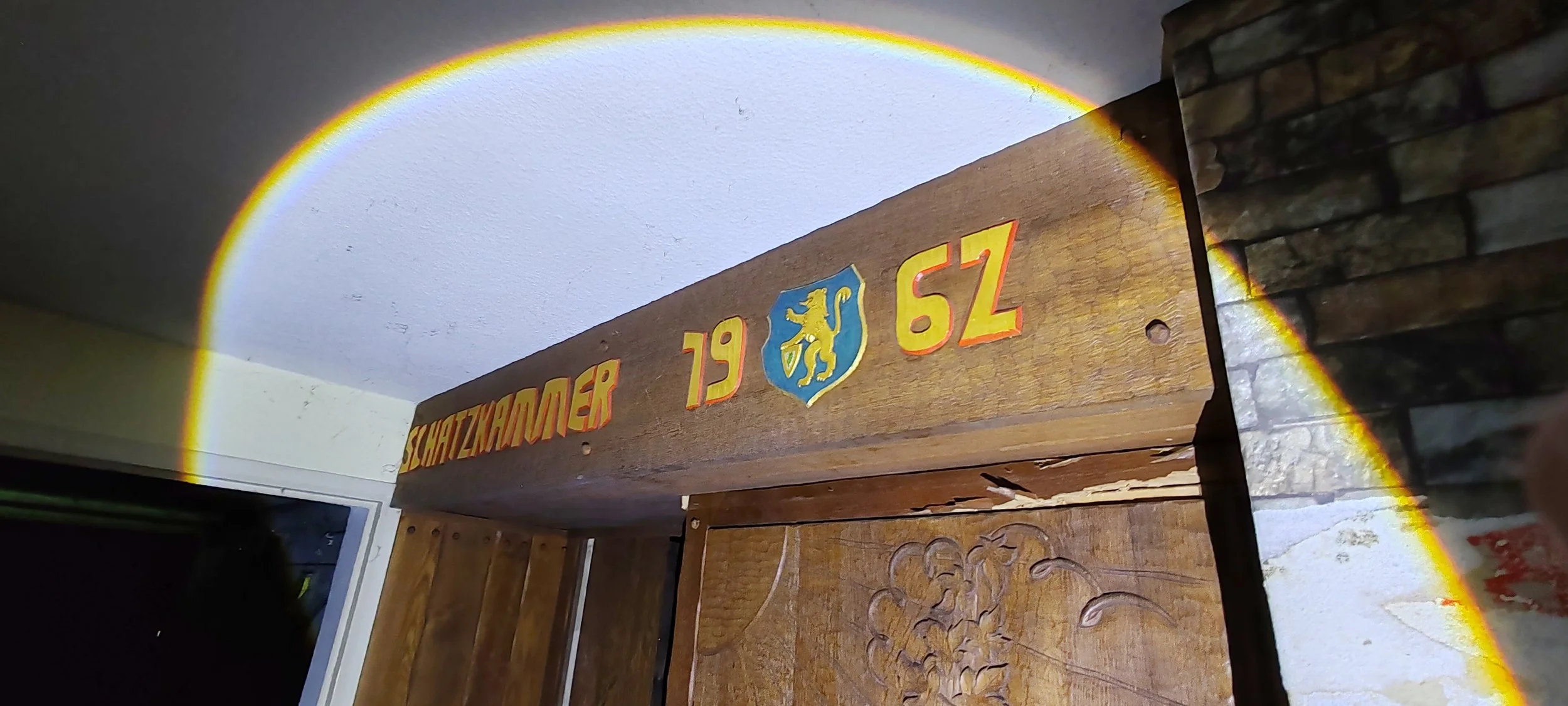 Holztafel mit Farben und Wappen, darunter die Aufschrift 'Schatzkammer 1962', neben einer Steinswand.