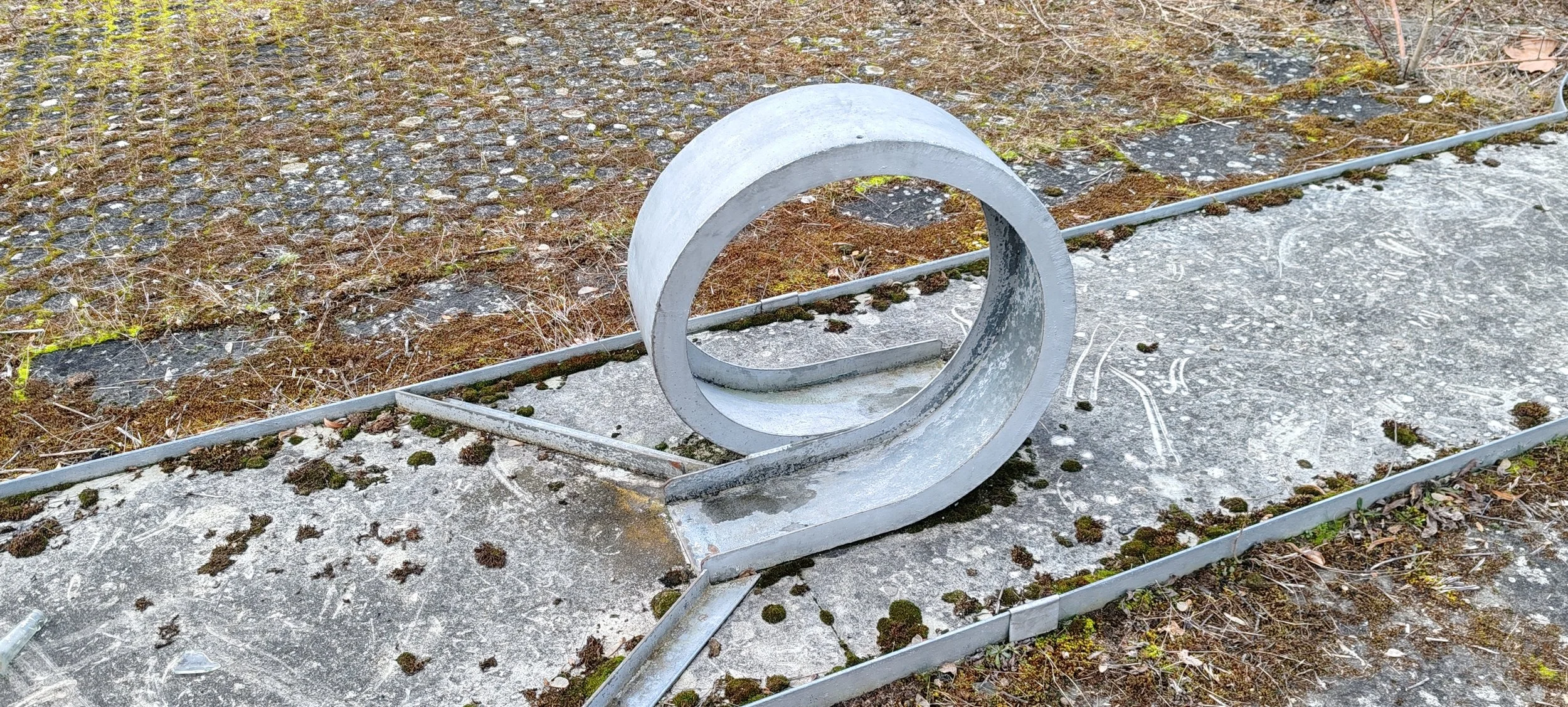 Ein spiralförmiges, silbernes Metallkunstwerk liegt auf dem Boden im Freien, umgeben von Moos, Erde und einer betonierten Fläche.