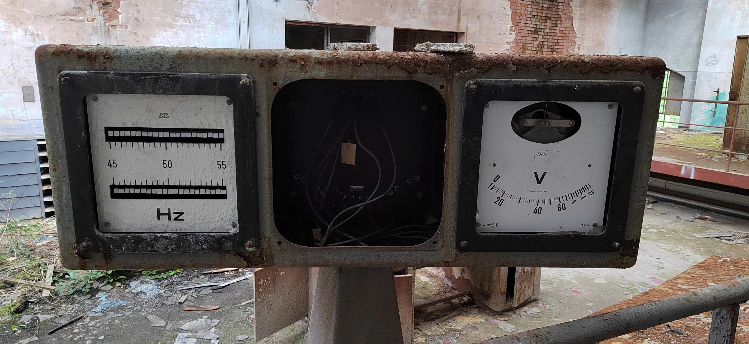 Ein alter, verrosteter Messgerät in einer verlassenen, verfallenen Industriehalle; es zeigt die Frequenz in Hertz, eine offene Stelle mit Drähten und Kabel, sowie eine Spannungsanzeige in Volt.
