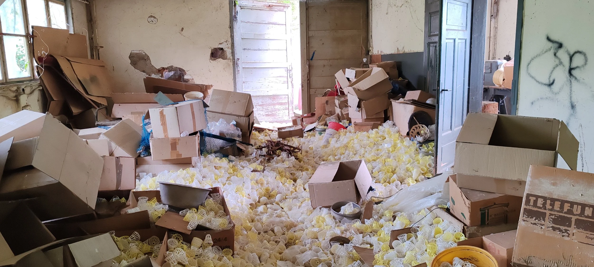 Der Raum ist voll mit zerknülltem und liegendem Verpackungsmaterial, Kartons, und anderem Müll. Die Wände sind beschädigt, und es gibt eine offene Tür, die nach draußen führt. Das Fenster auf der linken Seite lässt Licht in den Raum.