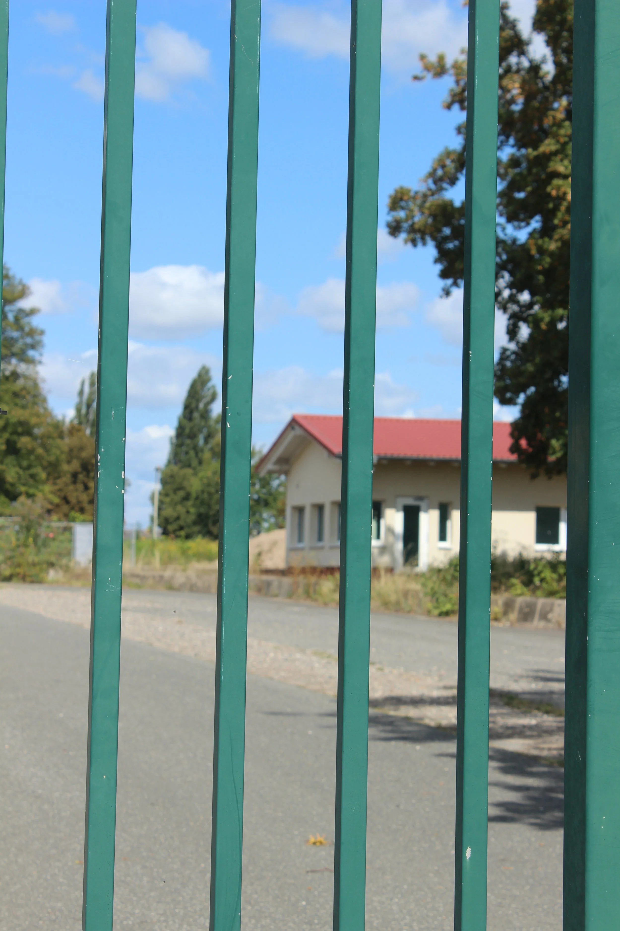 Blick durch eine Gitterzaun auf ein Haus mit rotem Dach und blauen Himmel mit Wolken.
