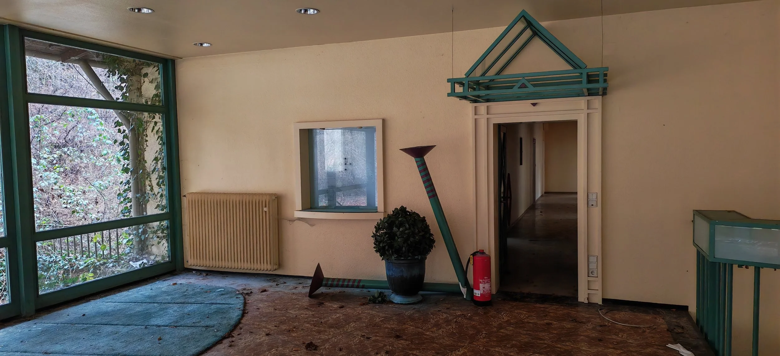 Innenraum mit großen Fenster, Blättern und Ästen draußen sichtbar, veralteter Heizung, kleine Fensteröffnung, Pflanzen, einen umgekippten Leuchter, Feuerlöscher, und nebenan abgesperrtes Zimmer. Der Raum wirkt ungepflegt.