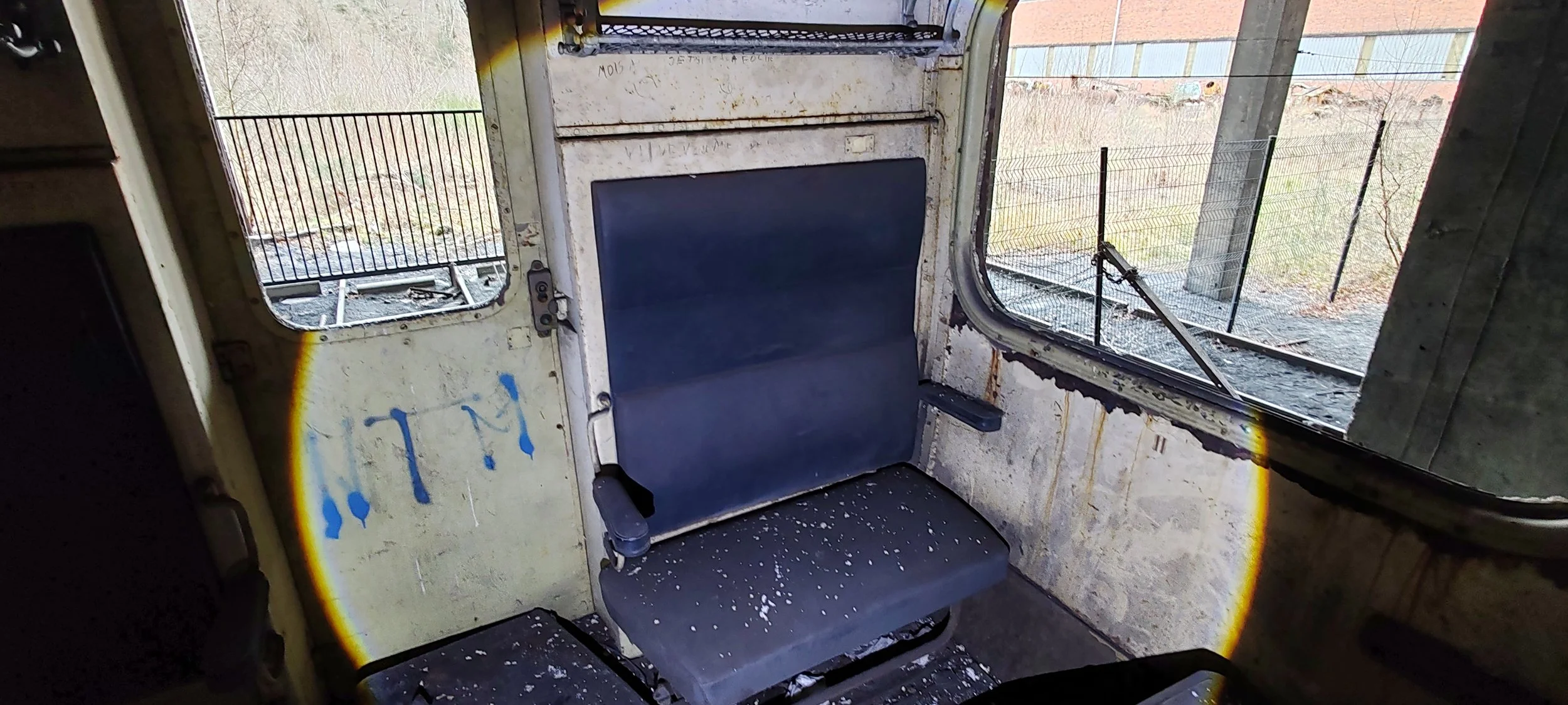 Verlassenes, verrostetes Inneres eines alten, kaputten Fahrzeugs mit Sitz und Fenster, draußen sichtbarer Zaun und wild bewachsene Flächen.