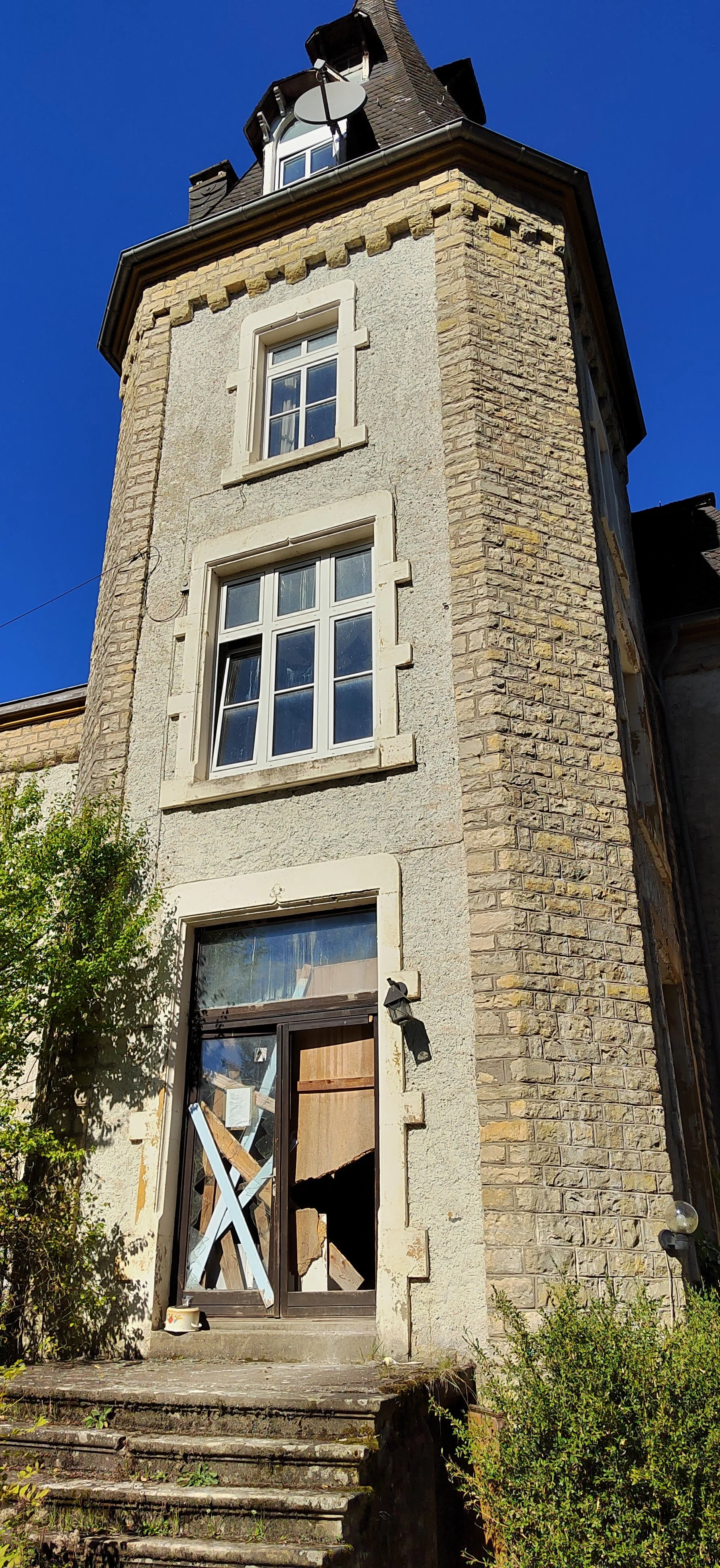 Ein altes, zweistöckiges Gebäude mit einer Tür, die durch Bauarbeiten beschädigt ist, und mehreren Fenstern, darunter ein kleiner Dachbodenfenster und ein großes Fenster im Erdgeschoss, bei sonnigem Himmel.