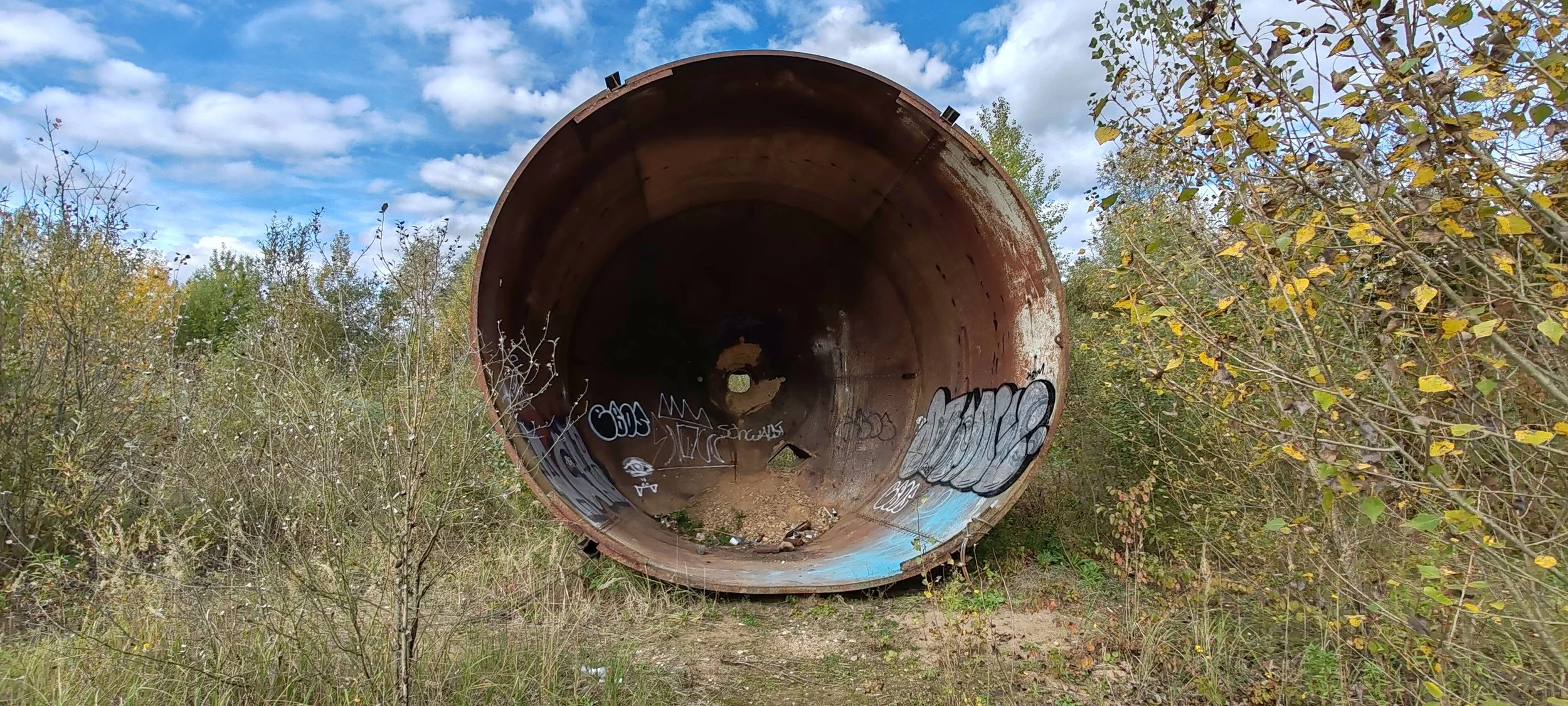 Großer, verrosteter, verwaister Metallrohr in einem überwachsenen, bewaldeten Gebiet mit Graffiti auf der Innenwand. Der Himmel ist teils bewölkt.