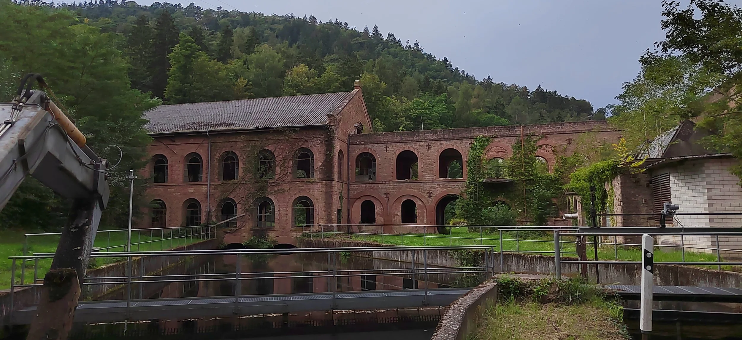 Historisches rotes Backsteingebäude mit vielen Bögen und einem Wasserkanal davor, umgeben von grünen Bäumen und hügeligem Wald. Auf der linken Seite ist ein Baukran sichtbar.