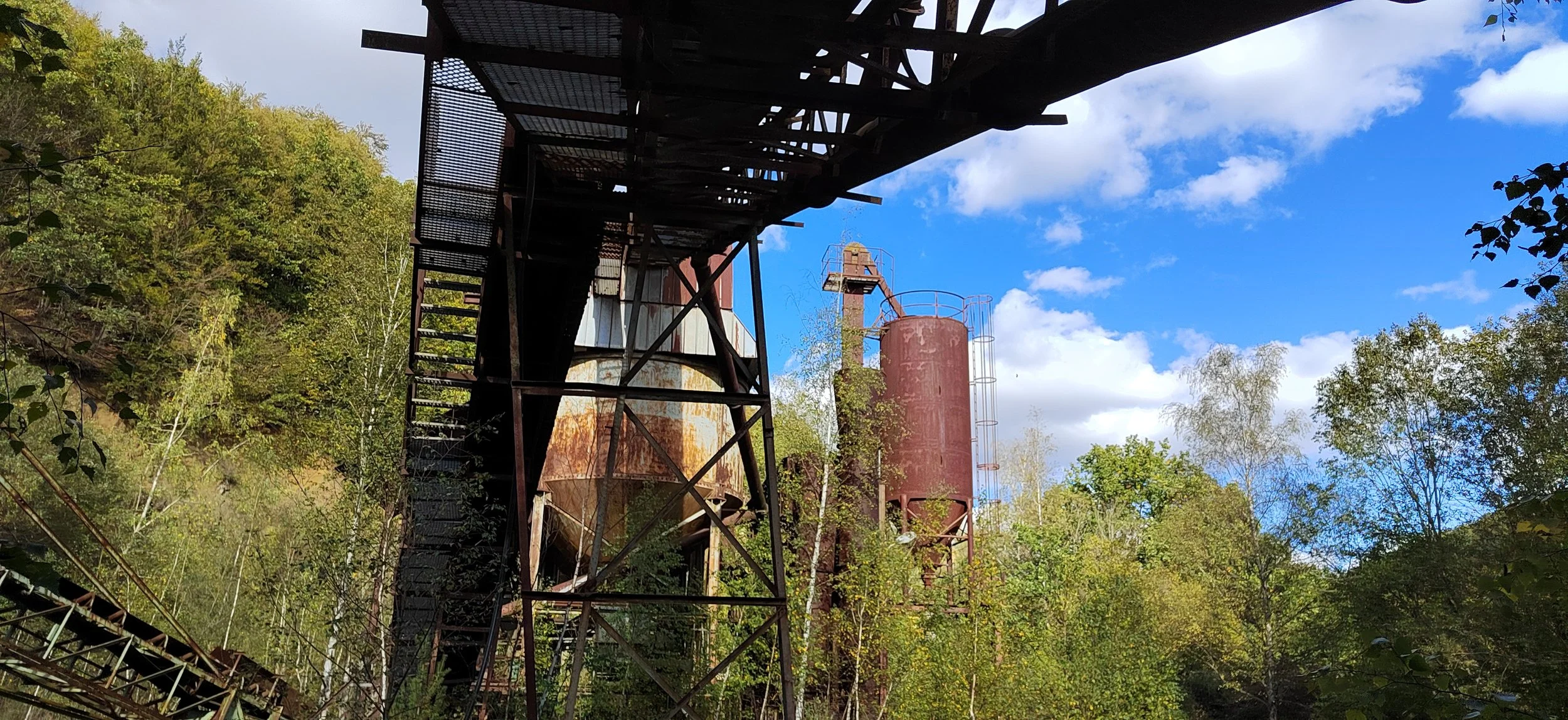 Verrostete alte Industrieanlagen inmitten eines Waldes bei blauem Himmel.