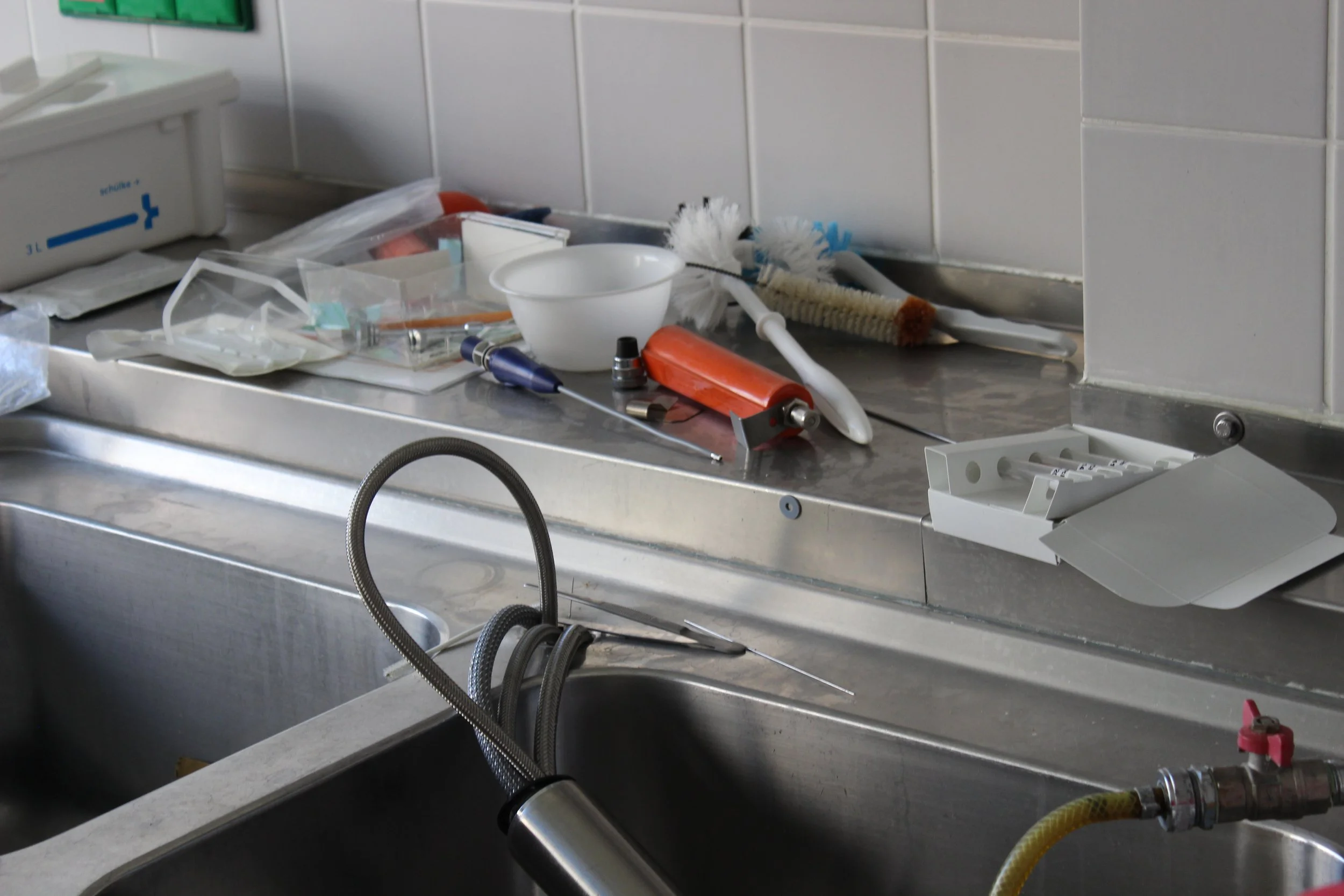 Ein verschmutzter Krankenhaus- oder Arztpraxis-Arbeitsbereich mit medizinischen Instrumenten, Schutzkappen, Schläuchen und Verpackungen auf einer Edelstahl-Arbeitsfläche.