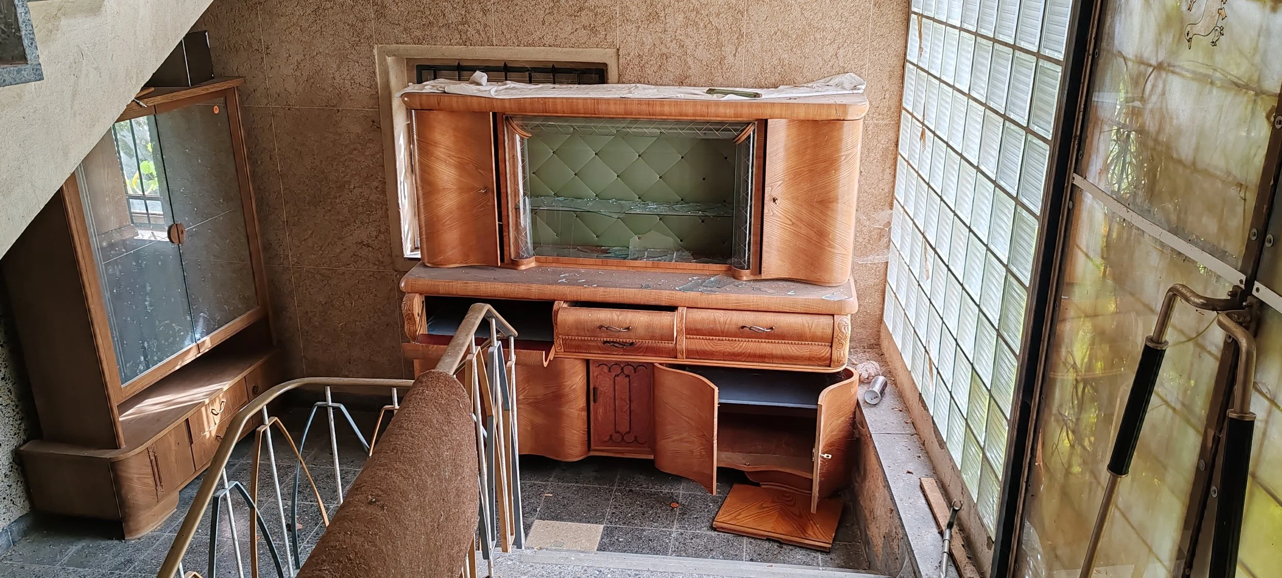 Ein alter, zerbrochener Holzschrank vor einer Ziegelwand, mit einem verbeulten Glasfenster im Hintergrund, auf einem Fliesenboden.