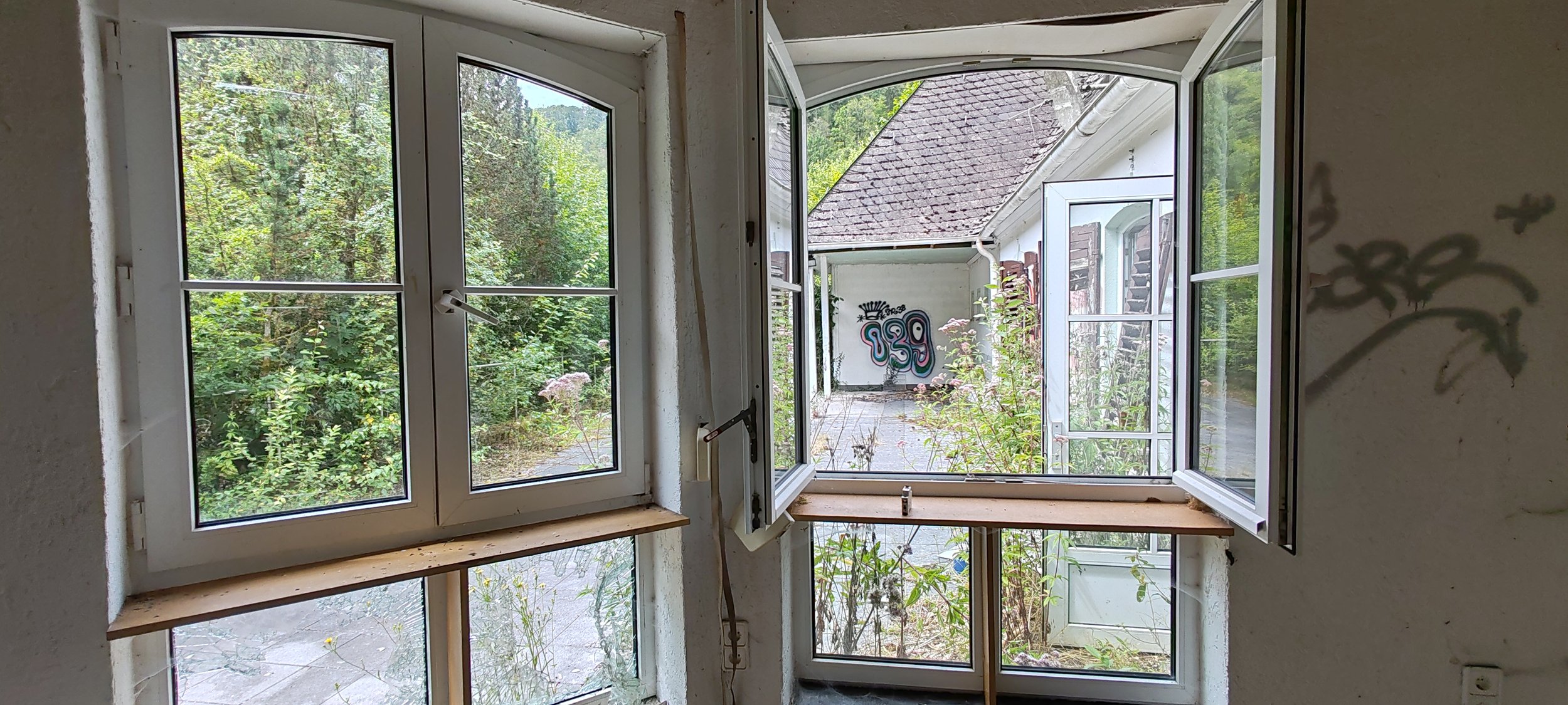 Offener Fensterblick nach draußen auf einen bewachsenen Garten mit Graffiti an der Wand auf der Terrasse, alte Häuser in der Nähe.