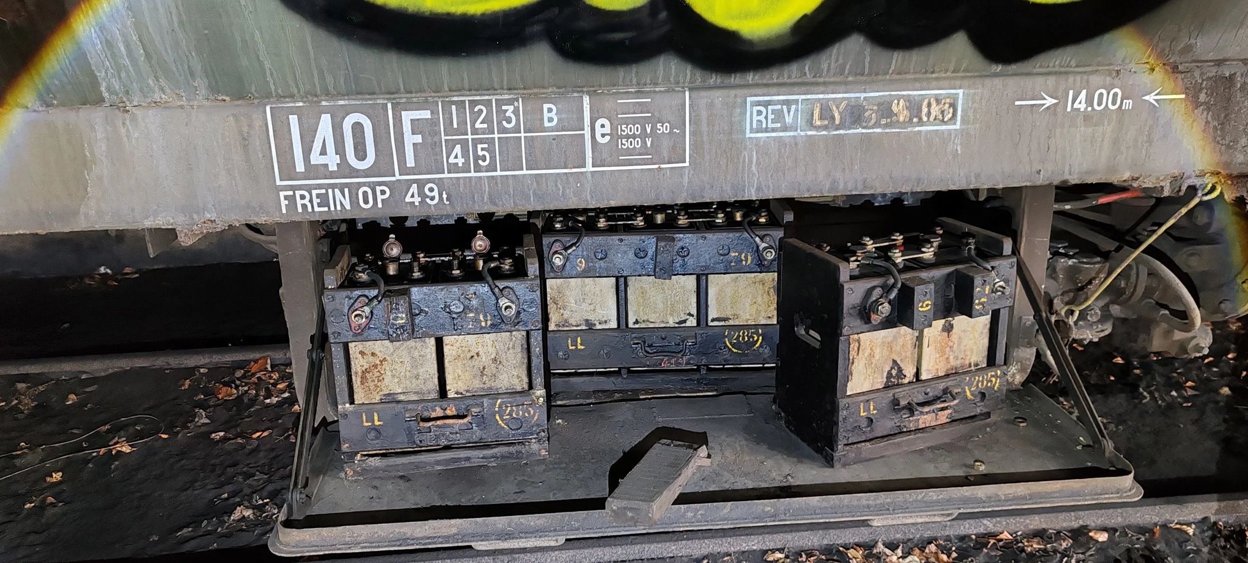 Schienenfahrzeug mit Beschriftungen und technischen Komponenten, einschließlich Gewicht und Spannung, auf der Unterseite. Der Untergrund besteht aus Kies und Laub.