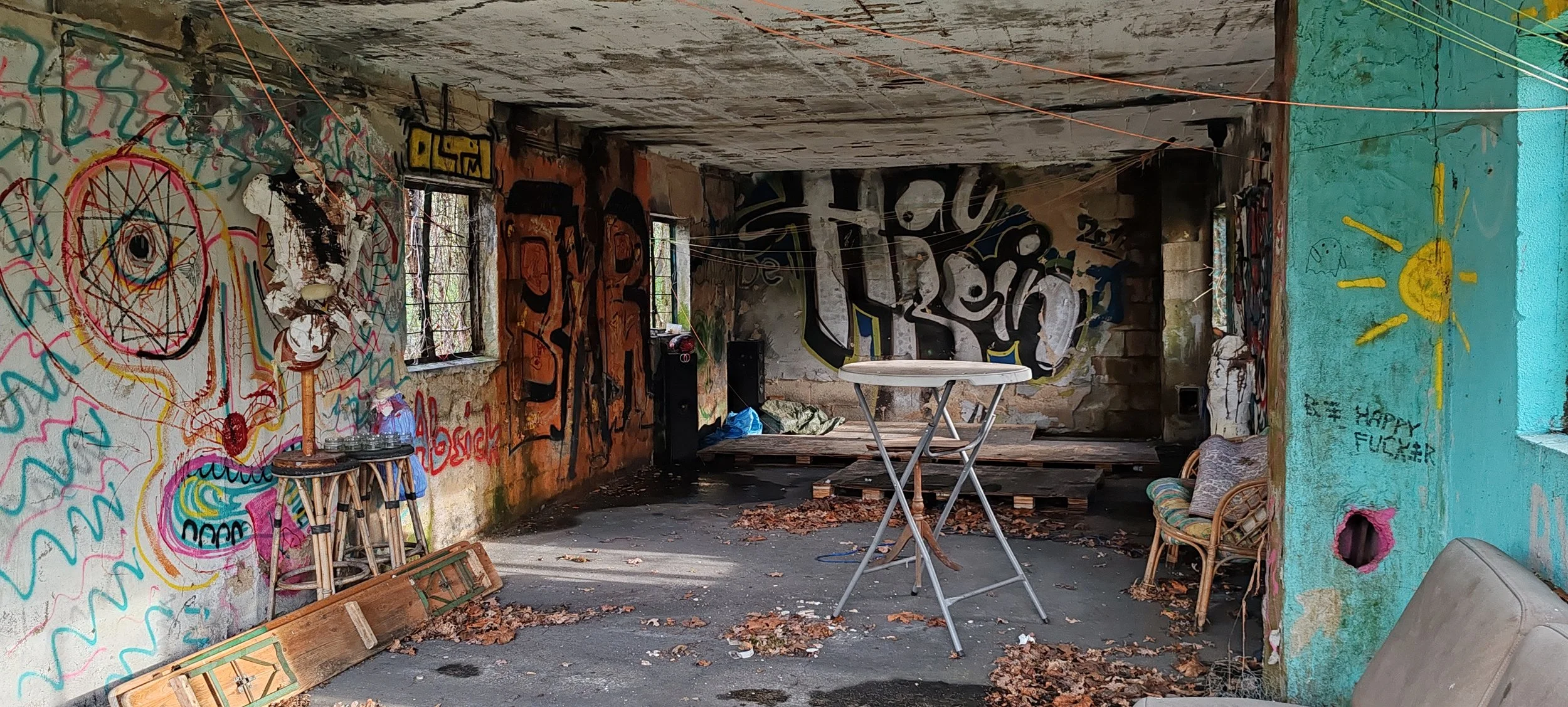 Verlassener, graffiti-überwachter Raum mit bekritzelten Wänden, leeren Wasserflaschen auf einem kleinen Tisch, zerfallenen Möbeln, Laub auf dem Boden und ungeordneter Umgebung.