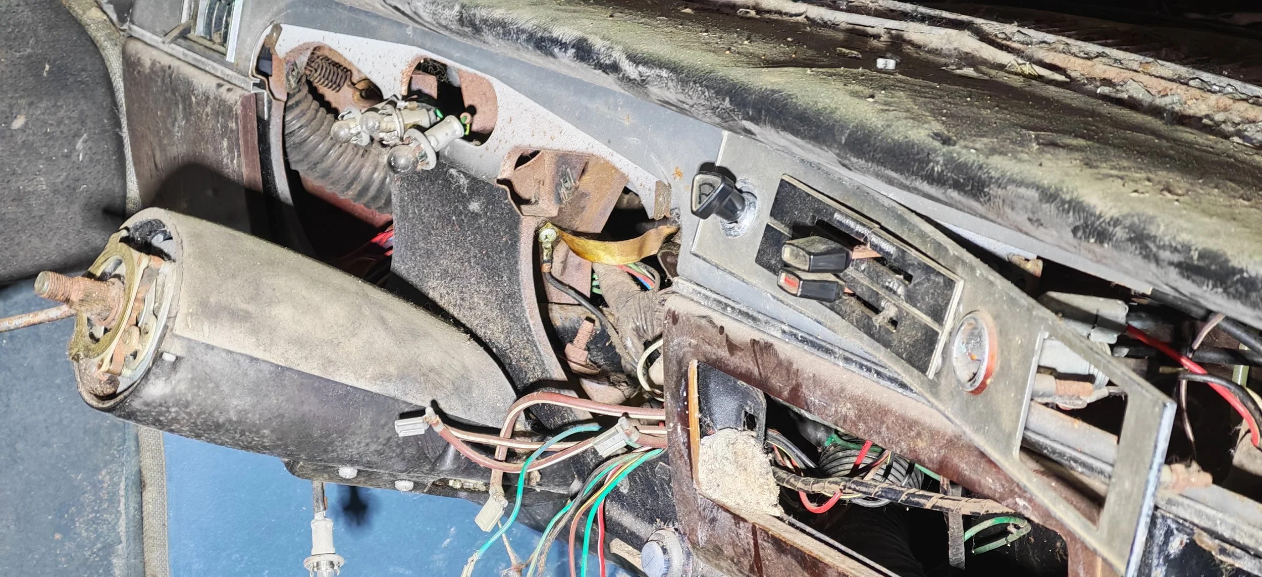 Das Bild zeigt das Innere eines alten, rostigen Fahrzeugs, einschließlich eines beschädigten Chassis, Kabel und elektrischer Komponenten, die sichtbar und teilweise verrostet sind.