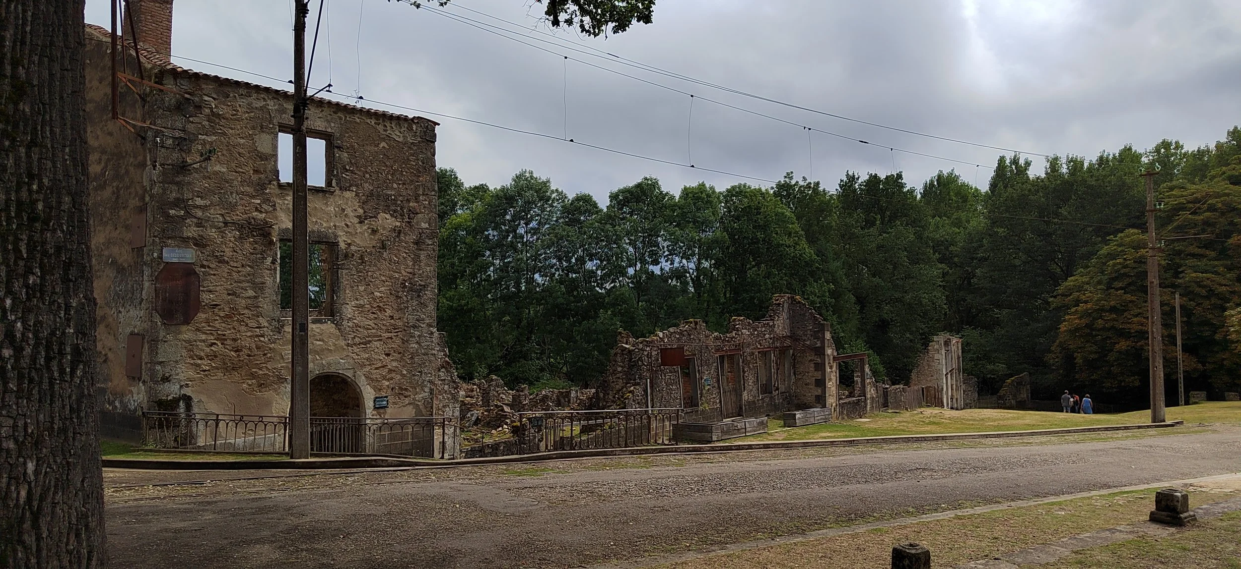 Ruinen eines alten Gebäudes neben einem grünen, bewaldeten Hügel unter einem bewölkten Himmel, mit drei Personen im Hintergrund.