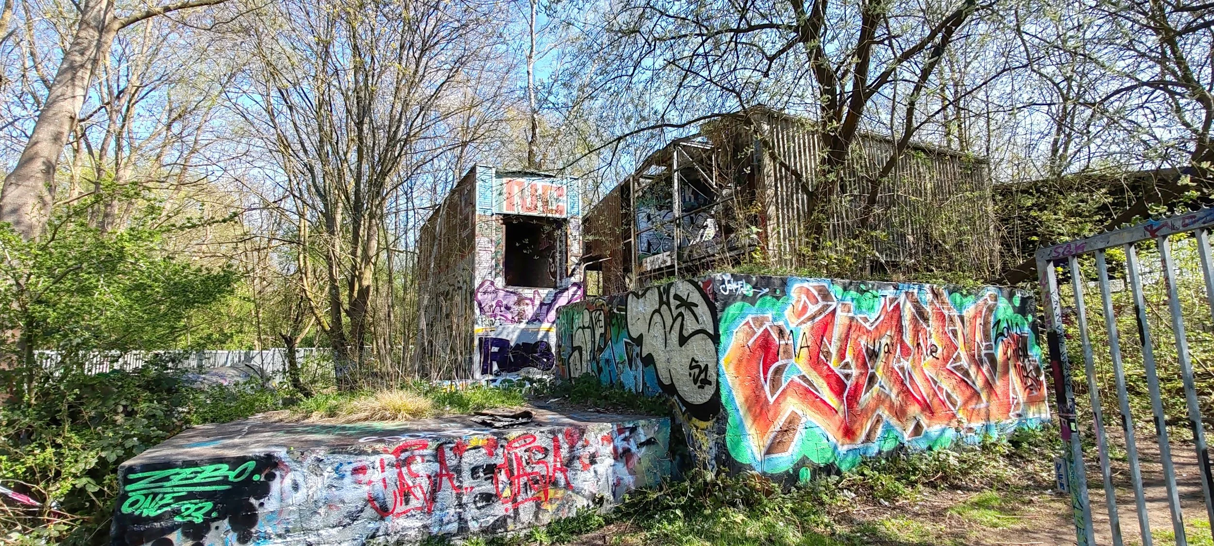 Verfallene, graffiti-bemalte Gebäude in einem Waldstück, umgeben von Büschen und Bäumen, mit einem Zaun auf der rechten Seite.