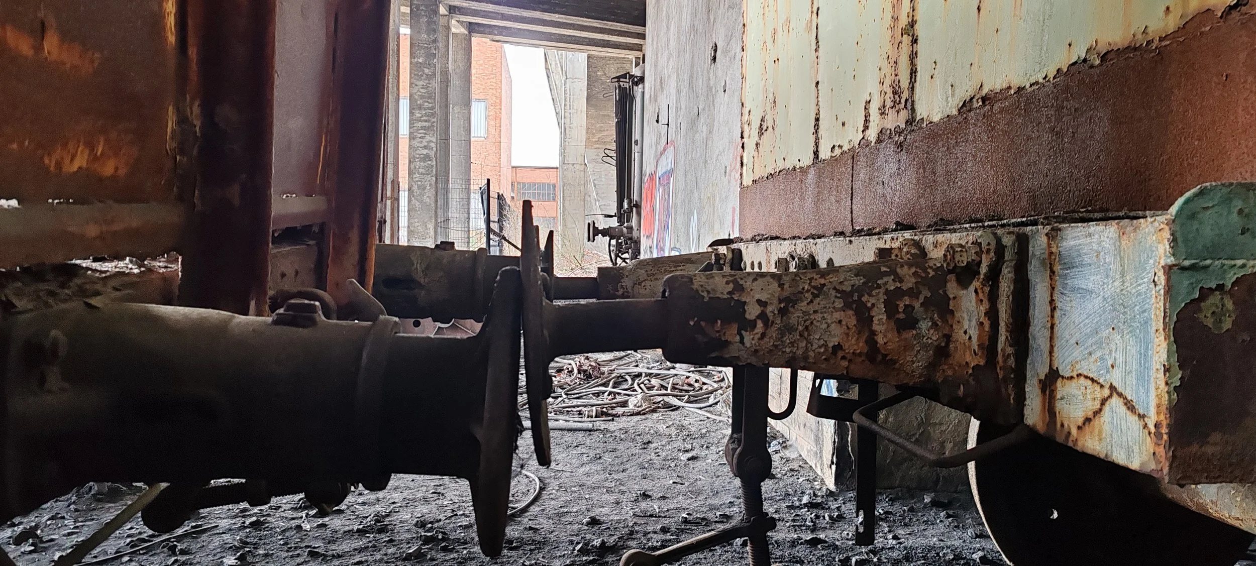 Blick unter eine alte, rostige, grüne Eisenbahnbrücke auf eine verlassene Bahnstrecke mit Streugut. Im Hintergrund sind moderne Gebäude sichtbar.