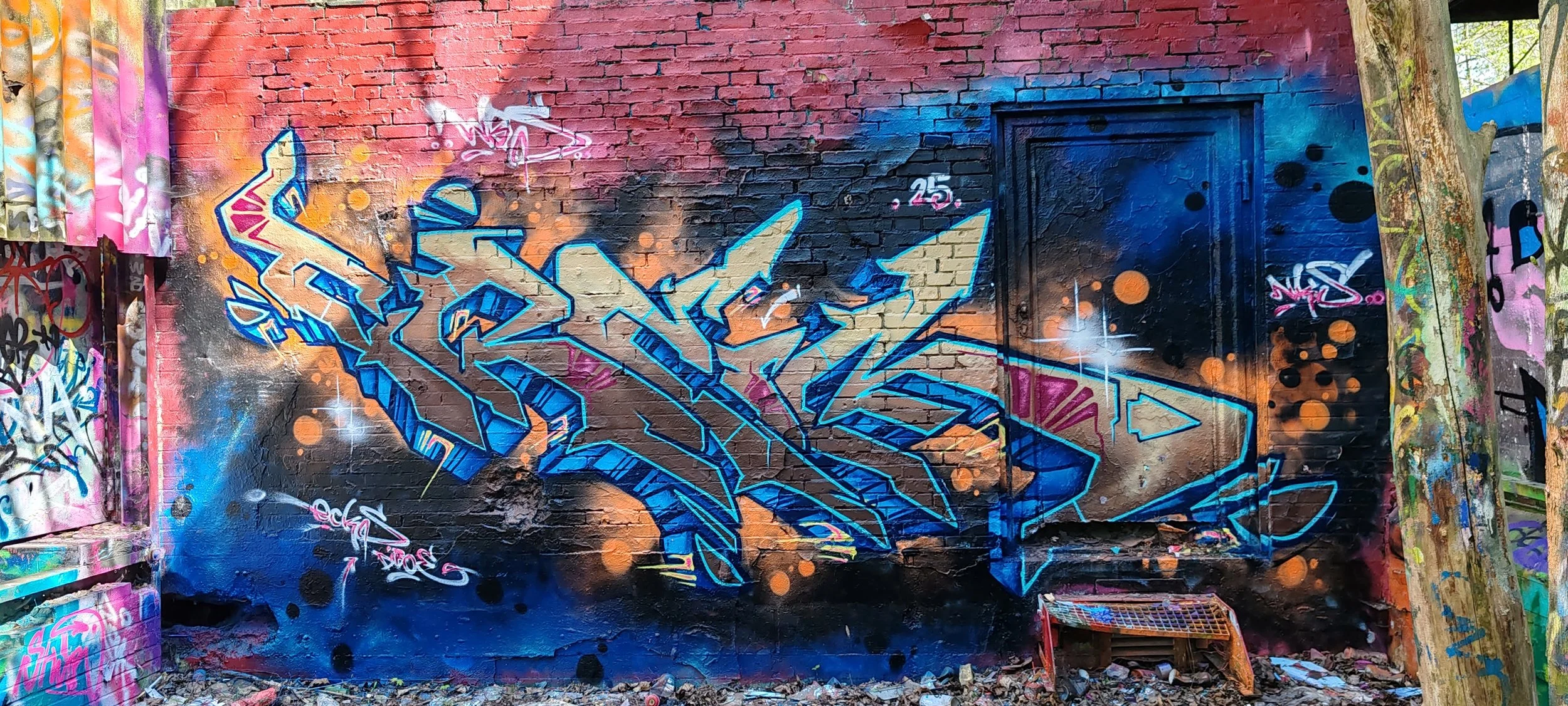 Graffiti-Kunstwerk an einer Wand mit bunten Farben, blauen, schwarzen, rosa und orange Akzenten, und einem grünen Baum auf der rechten Seite.