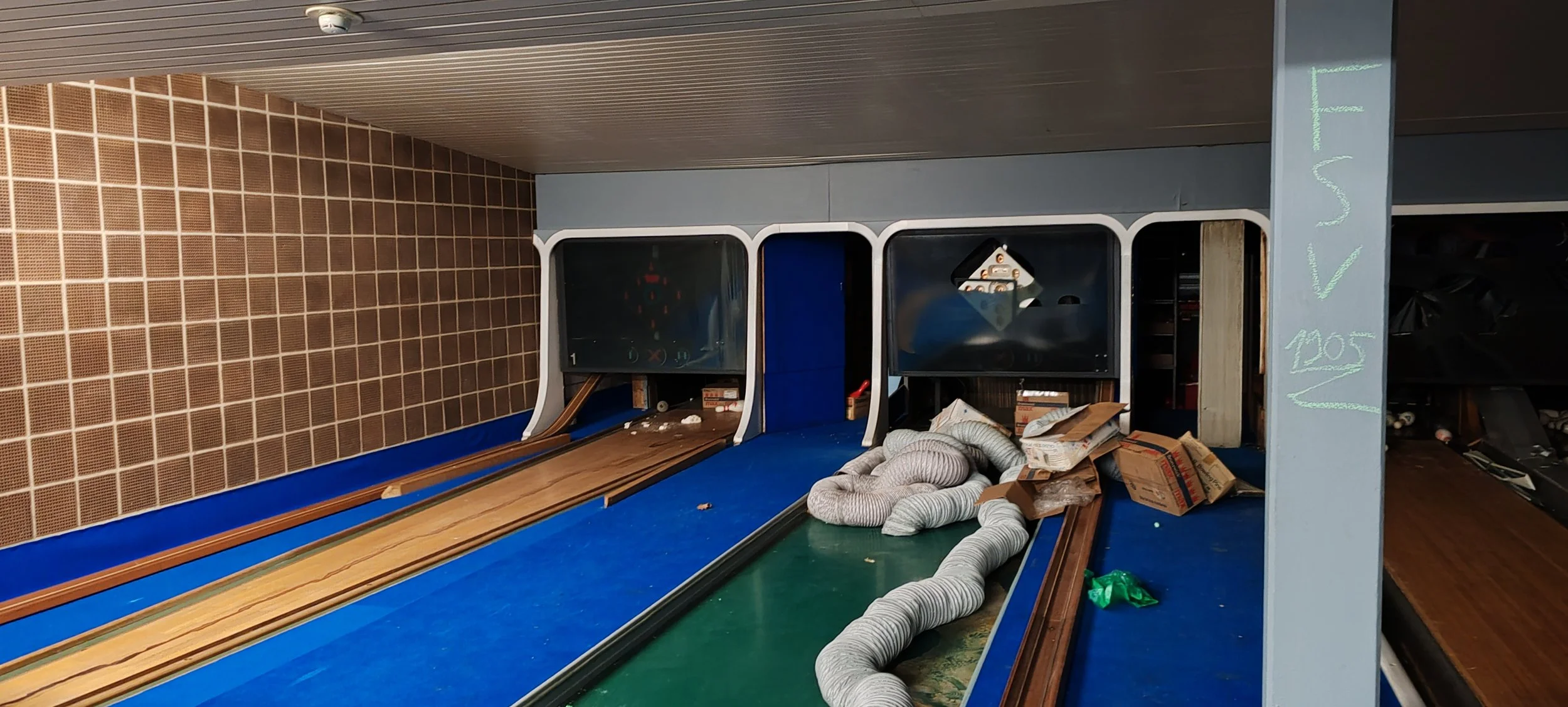 Baugleiche, alte Bahn mit Schienen, die vor der Fertigstellung steht, in einem Raum mit brauner Fliesenwand und grauem Deckenpaneel, neben einem blauen und schwarzen Kasten mit Holzteilen und Verpackungen, die auf der Bahn liegen.