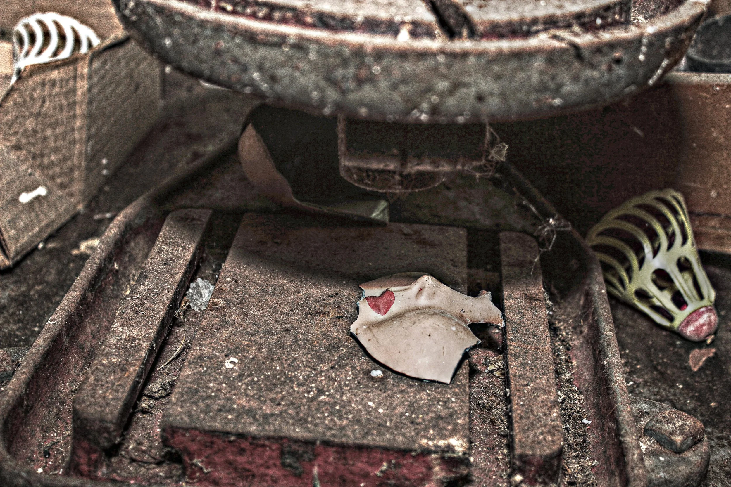 Alte, verrostete Schneidemaschine mit verstreuter Keramik auf der Oberfläche, im Hintergrund eine dekorative Schleifkugel, alles sichtbar in einem staubigen, verfallenen Raum.