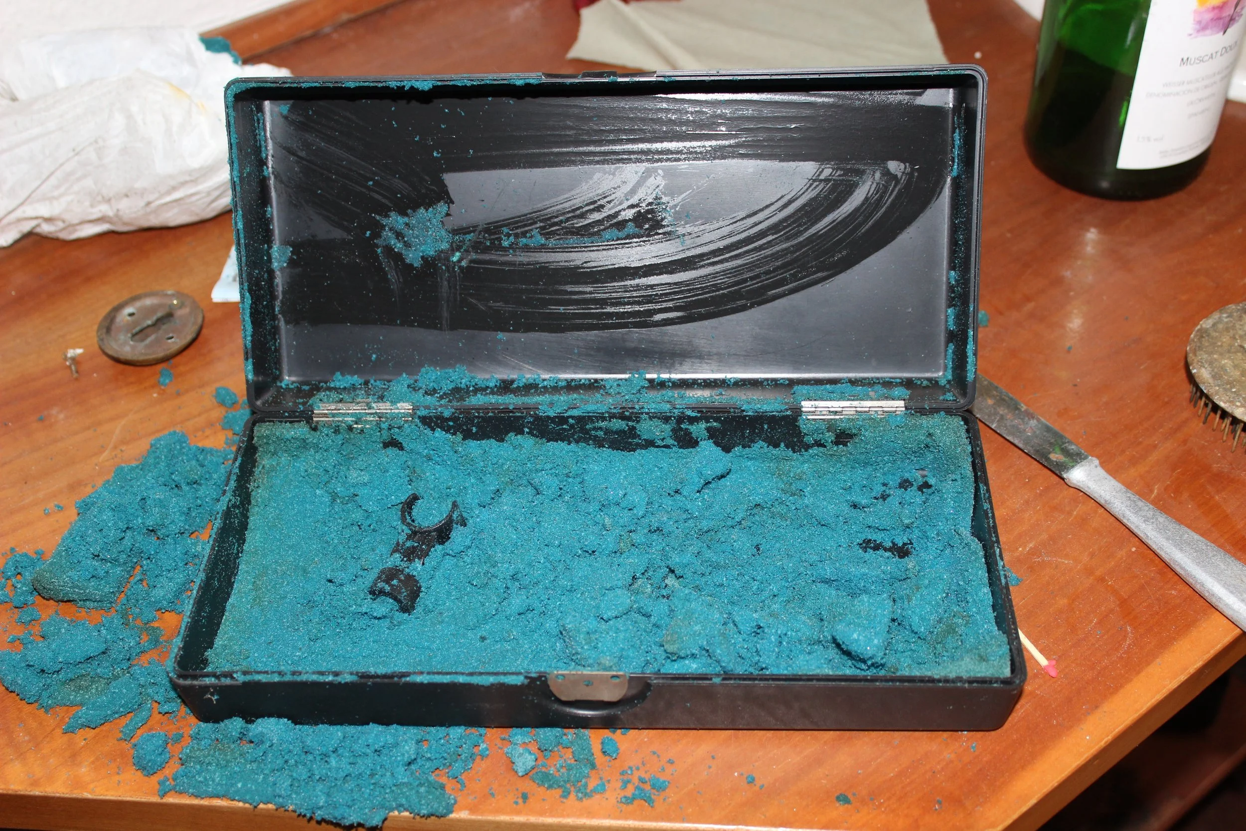 Aufgeklappte Brillenetui mit blauer Substanz und Werkzeug, umgeben von Verschmutzung auf einem Holztisch.