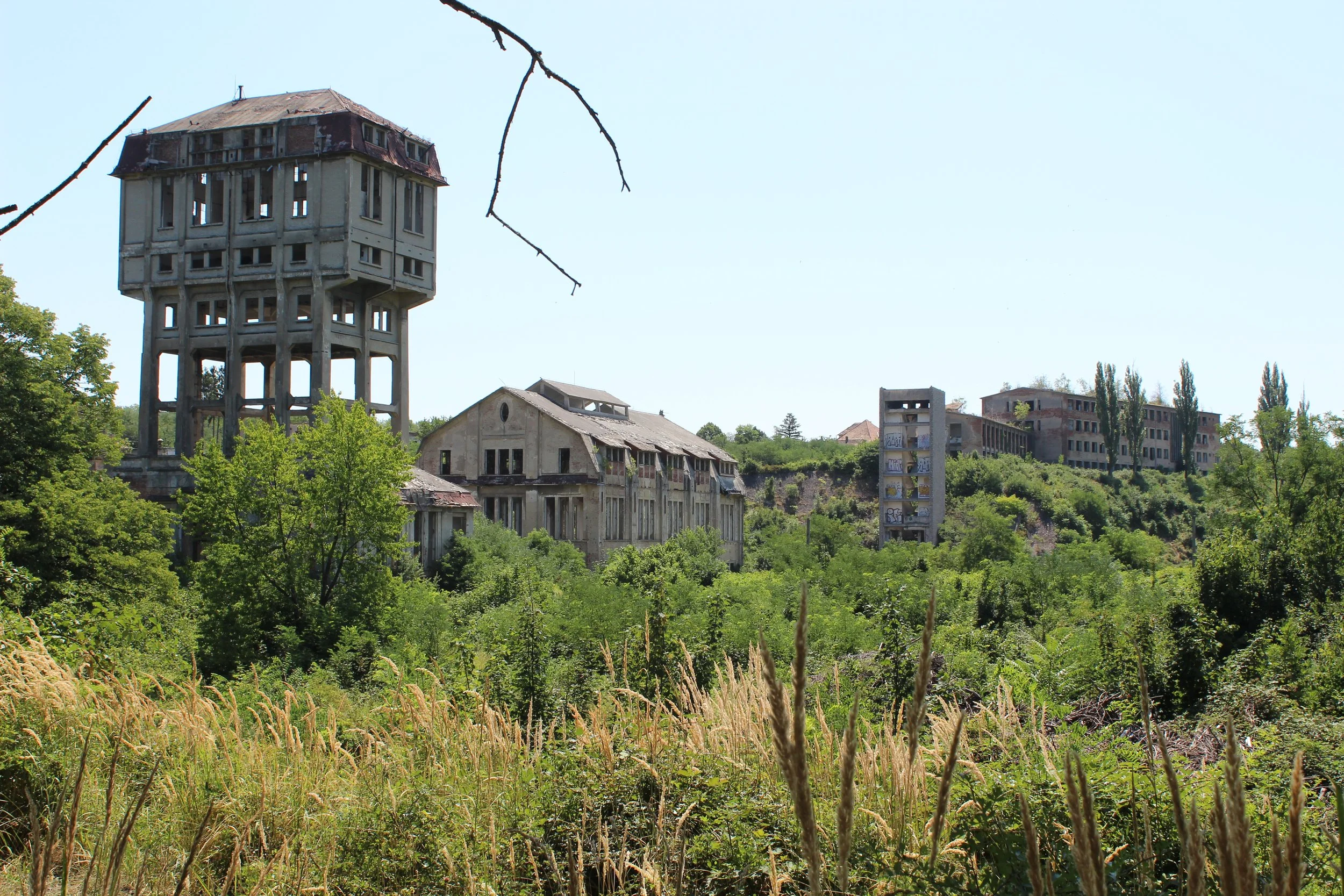 Verlassene und verfallene Gebäude in einer grünen, bewachsenen Umgebung, darunter ein Wasserreservoir auf Säulen, mehrere Häuser und Hochhäuser im Hintergrund, bei sonnigem Himmel.