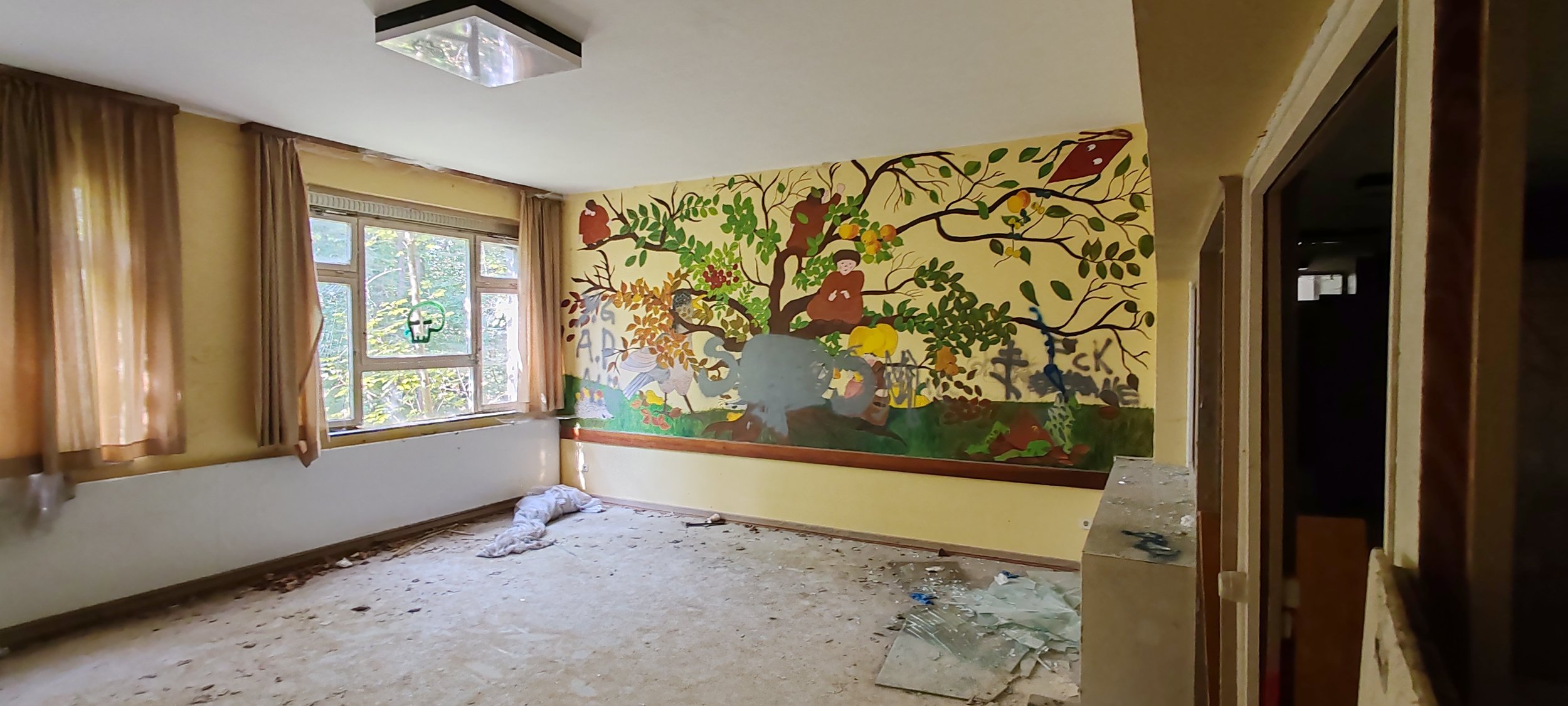 Ein leerer Raum mit buntem Wandgemälde eines Baumes mit Tieren und Menschen, große Fenster mit beige Vorhängen, unordentlicher Boden mit Müll