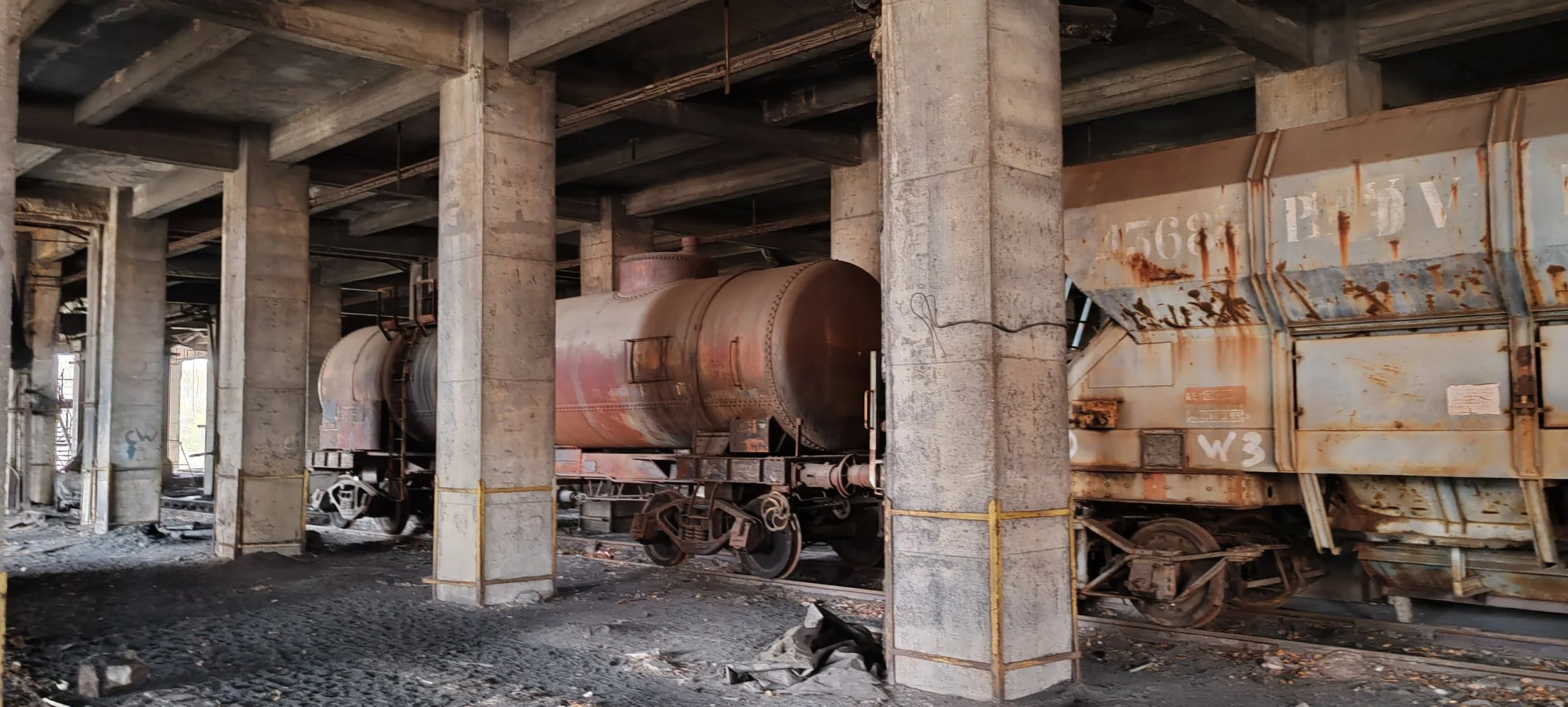 Alte, verrostete Eisenbahnwagen stehen in einer alten Lagerhalle mit Betonstützen und einer Holzbalkendecke, Staub und Schutt auf dem Boden.
