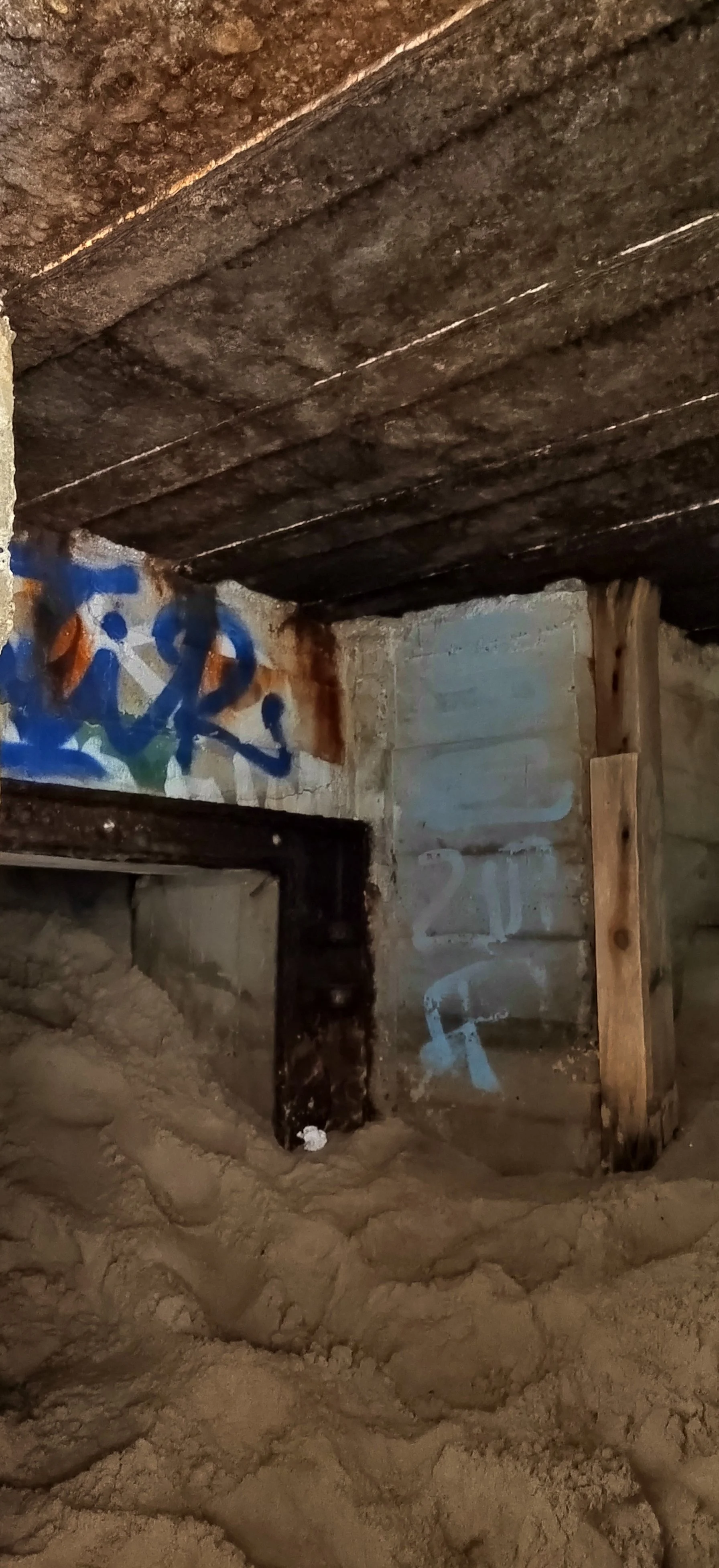 Blick unter einem alten Holz- und Ziegelboden, mit Graffiti an der Wand, Sandboden, Holzstützpfeiler und sichtbare Holzbalken