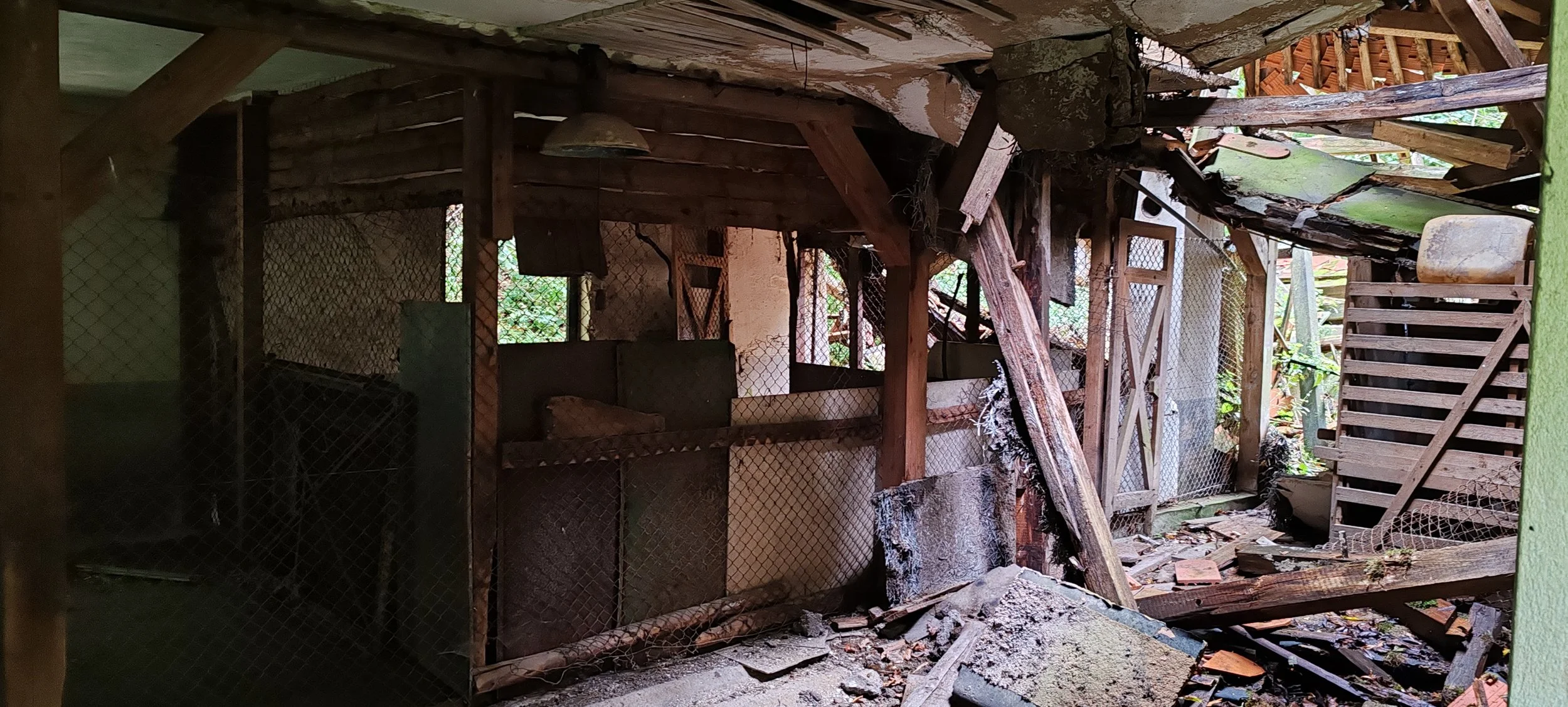 Ein verfallenes, zerstörtes Gebäude mit umgestürzten Holzteilen, zerbrochenen Steinen und zerfetzten Netzen, sichtbar durch offene Wände und Dachlücken, mit umgebenem Grün im Hintergrund.