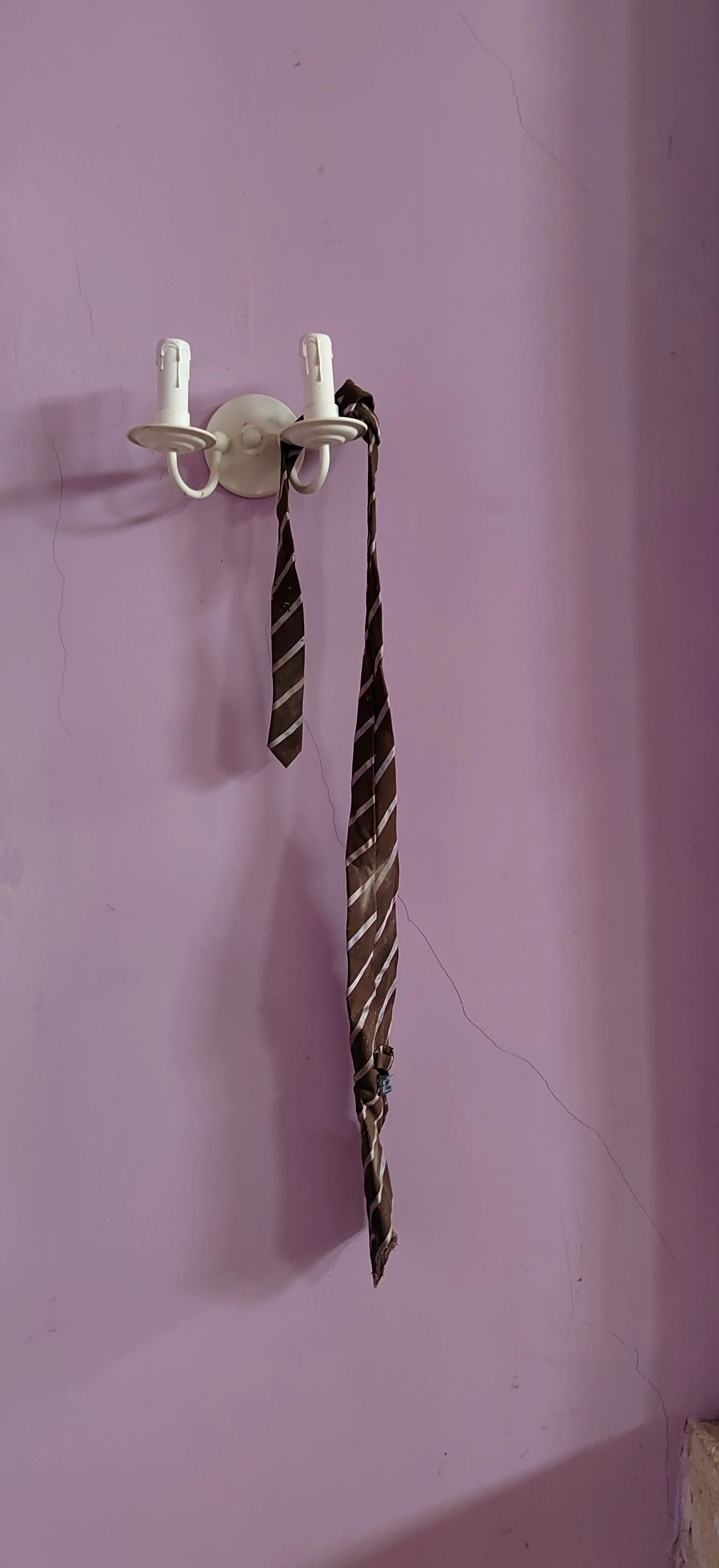 Ein Wandhaken mit zwei weißen Kerzenhaltern, an denen braune Krawatten hängen, an einer lilafarbenen Wand mit sichtbaren Rissen.