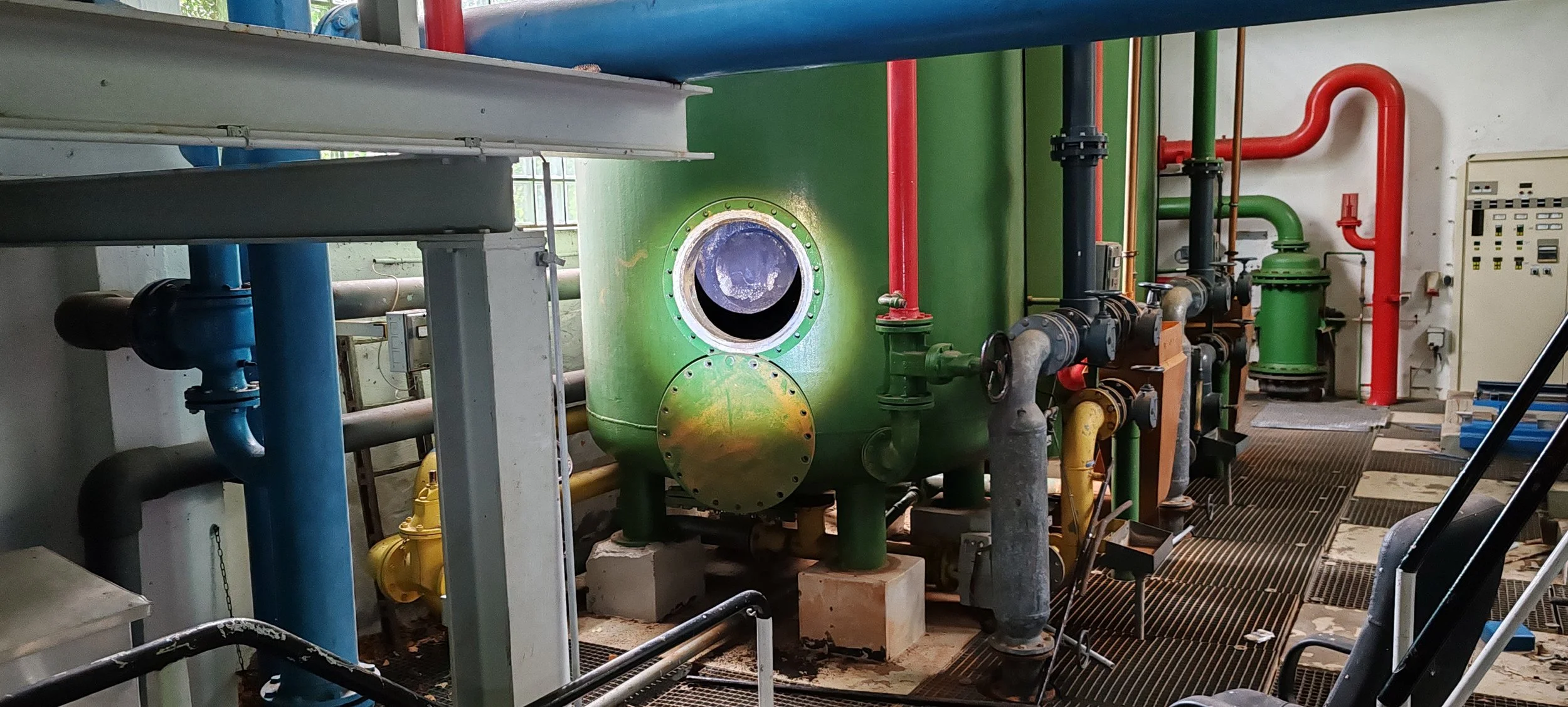 Blick auf eine technische Anlage mit grünen, blauen, roten und schwarzen Rohren, einer großen grünen Kessel und einem Schaltsystem im Hintergrund. Es ist wahrscheinlich in einer Industrie- oder Wartungsumgebung.