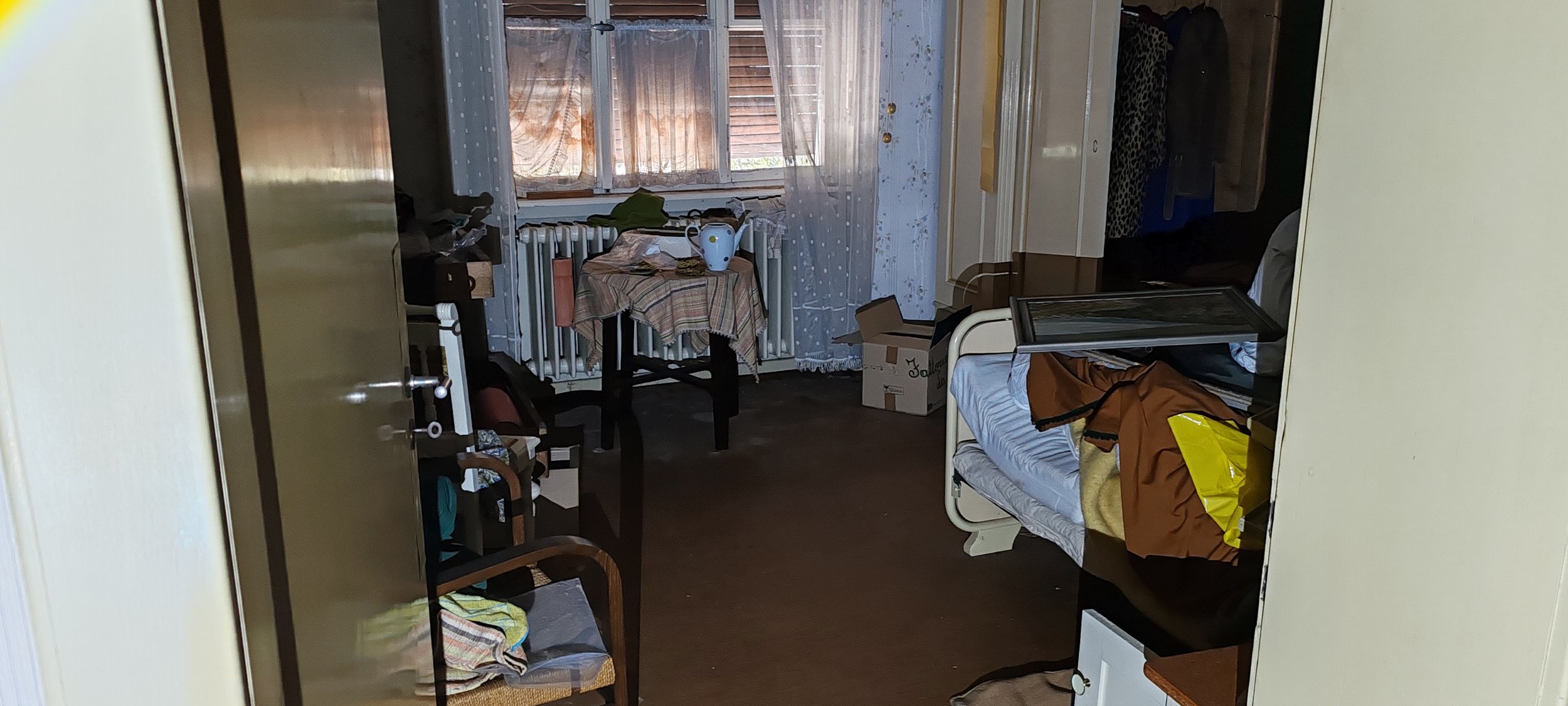 Ein unordentliches Schlafzimmer mit einem Bett, einem Nachttisch, einem Schreibtisch, einem Stuhl, einer Box, einem Wasserkocher auf einem Tisch, und einem Fenster mit Vorhängen, durch das Tageslicht scheint.