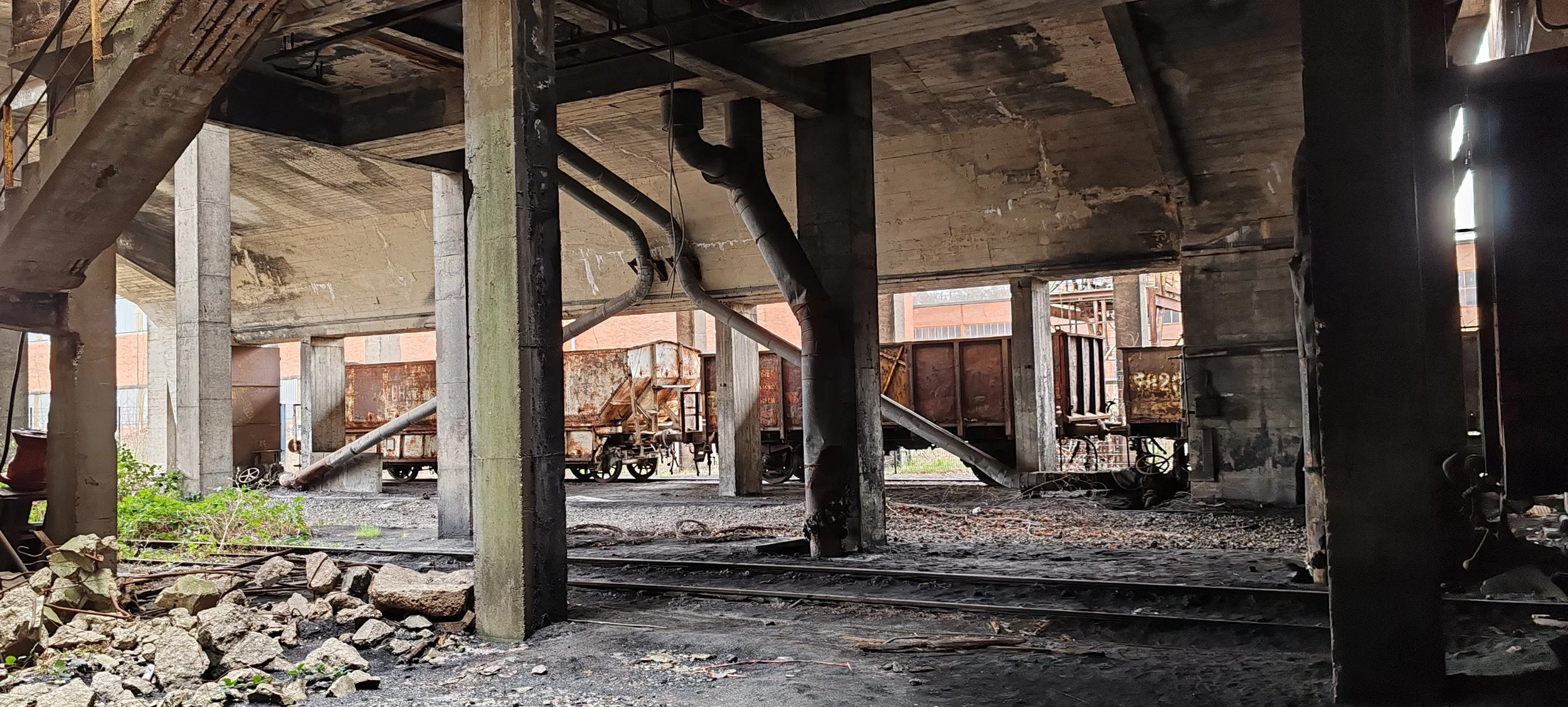 Verrostete Güterwagen unter einer Brücke auf einem alten Eisenbahngleis, mit Steinschutt und Pflanzen im Vordergrund.