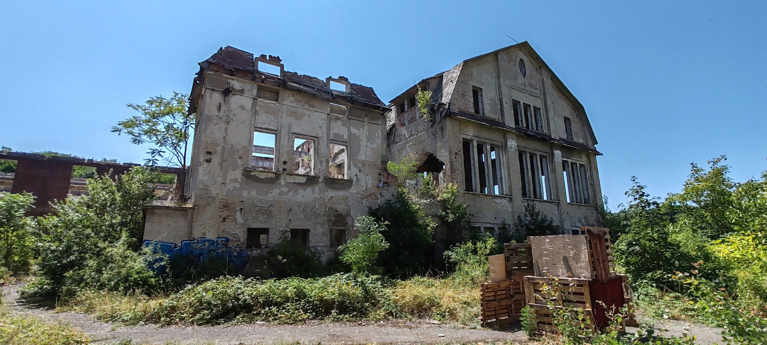 Verfallene, verlassene Villa mit beschädigtem Dach und zerbrochenen Fenstern, überwuchert von Pflanzen und Bauschutt im Vordergrund, gegen blauen Himmel.
