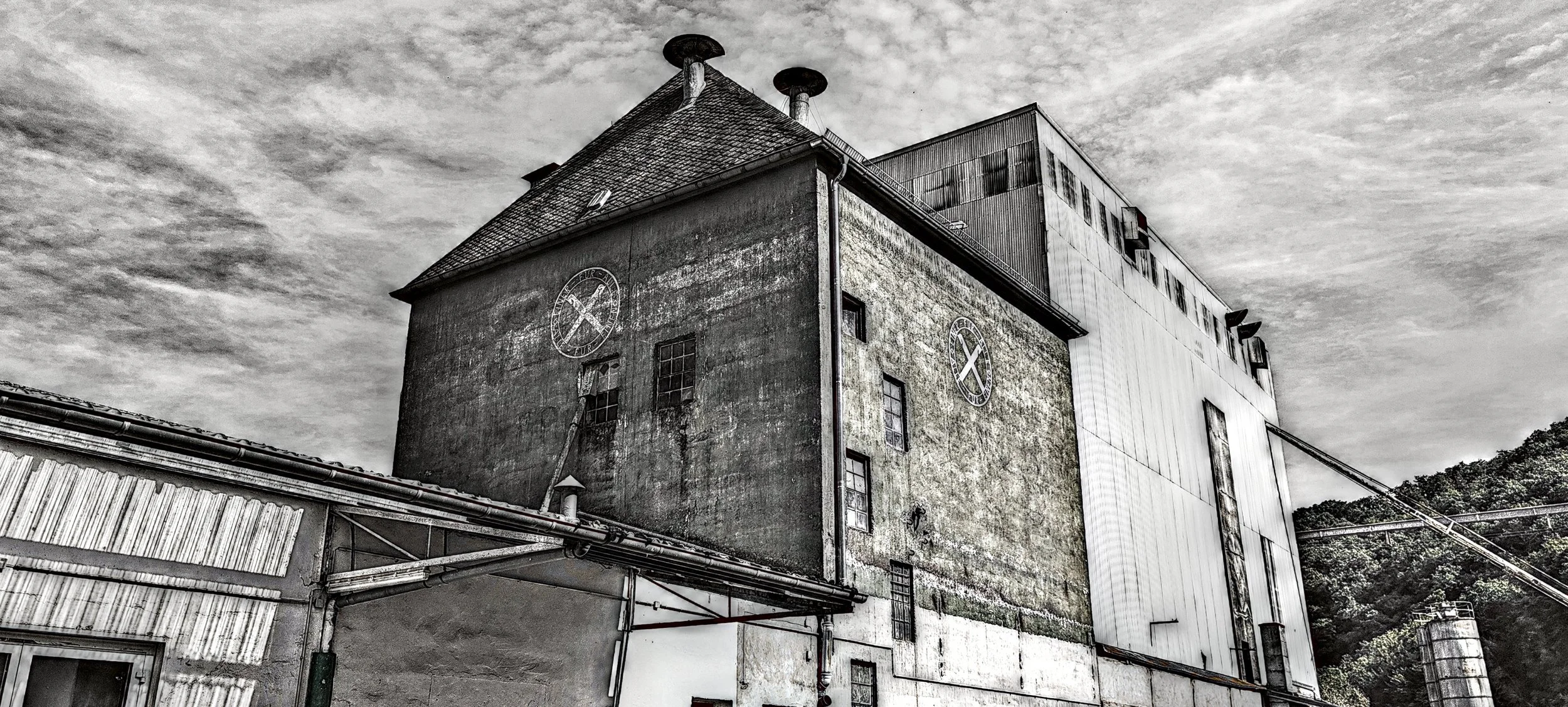 Ein altes Industriegebäude mit zwei großen Uhren an den Fassaden. Das Gebäude ist grau und zeigt Verfallsspuren, während im Hintergrund ein moderneres, weißes Gebäude mit Metallfassade zu sehen ist.