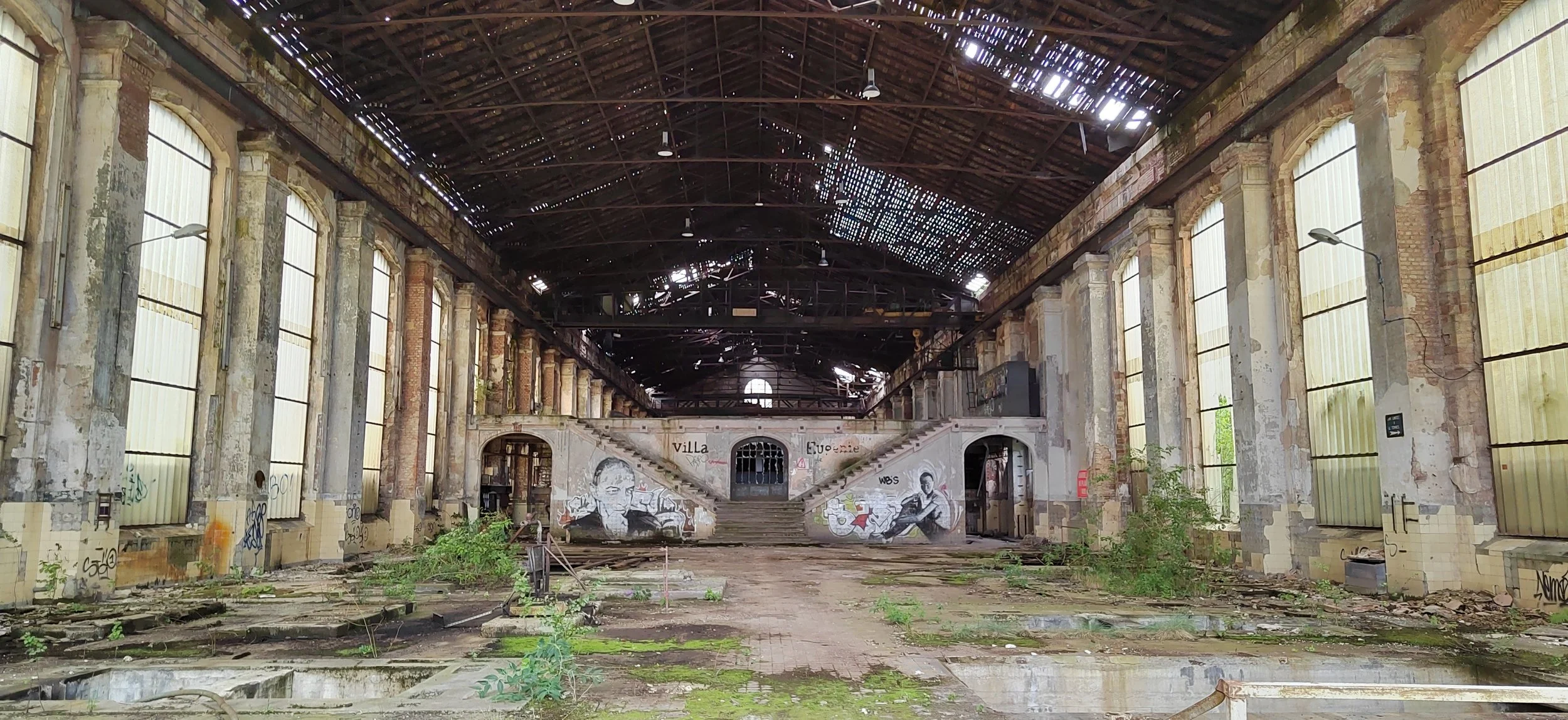 Innenansicht einer verlassenen, verfallenen Industriehalle mit graffiti-besprühter Wand, überwucherten Böden und einem zerbrochenen Dach, durch das Licht einfällt.