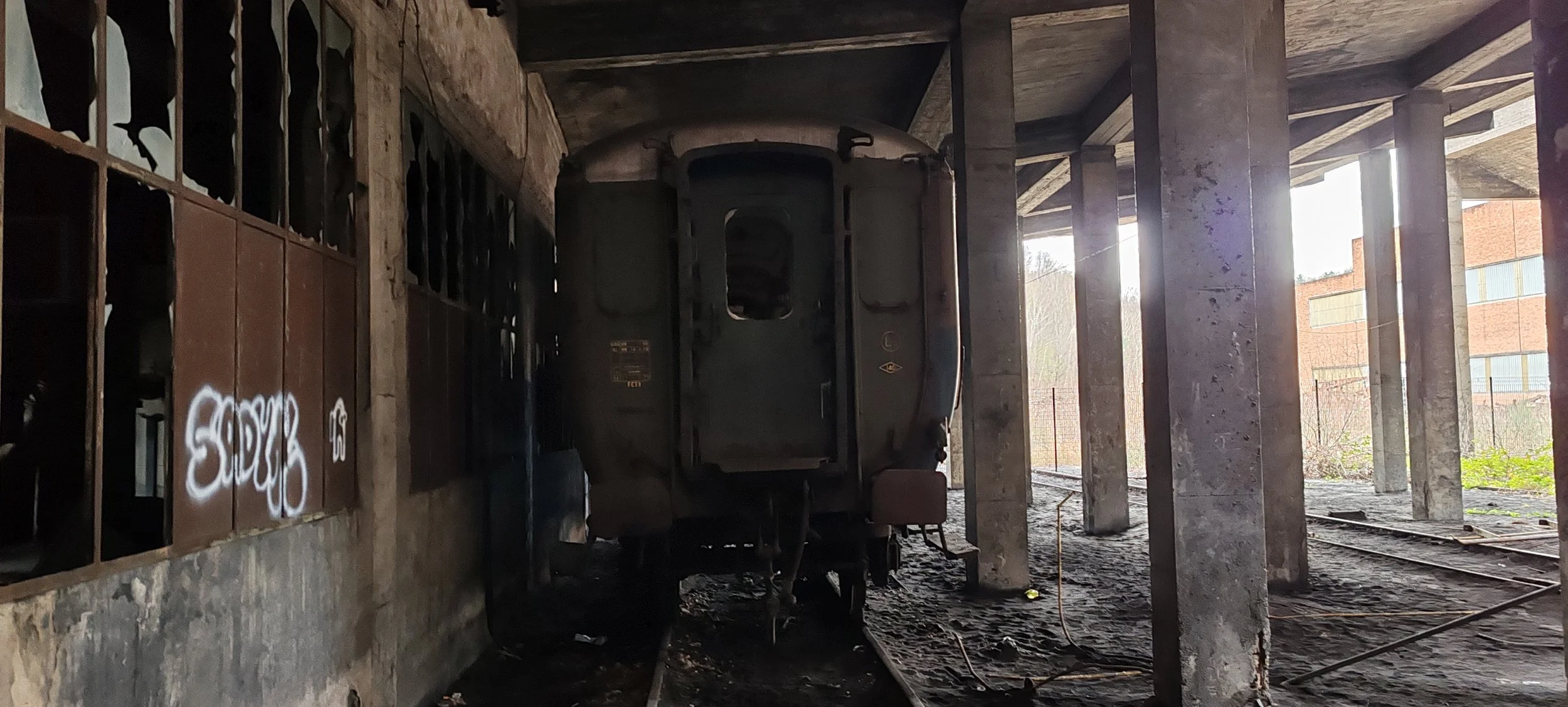 Verrosteter, alter Zug in einer dunklen, abgerockten Abstellhalle mit Graffiti an der Wand und Sonnenlicht, das durch die Fenster auf die Seite des Zuges fällt.