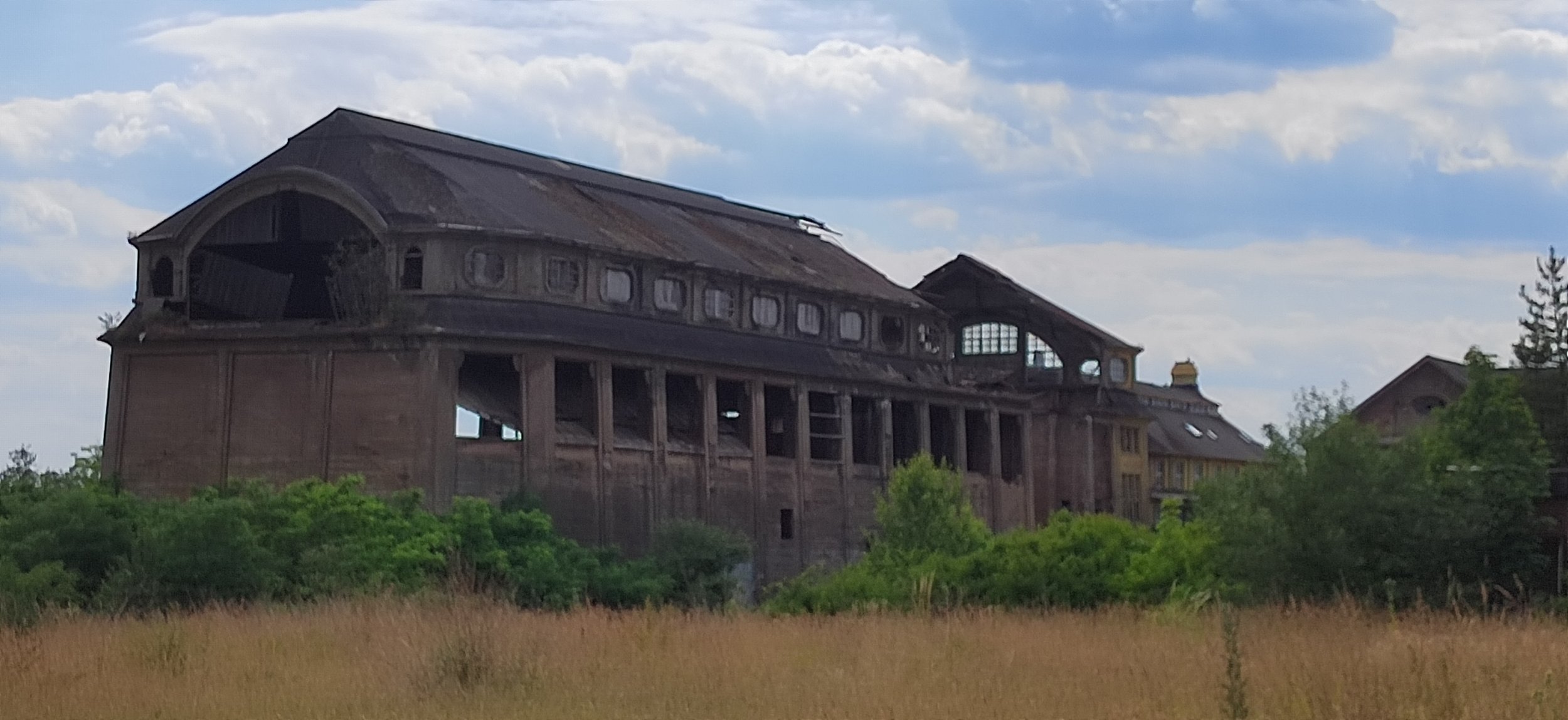 Verfallenes, altes Gebäude inmitten grüner Natur, unter einem bewölkten Himmel.