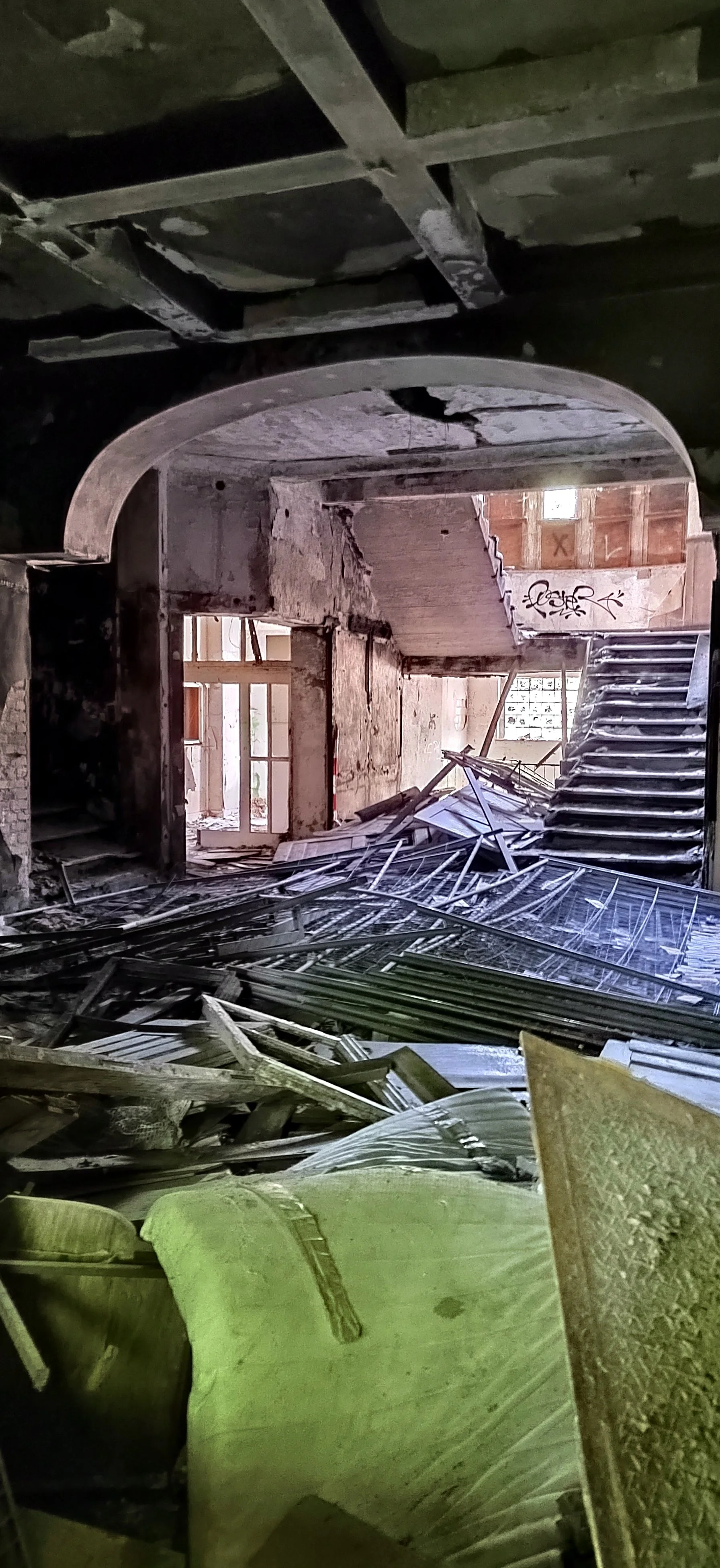 Ein verfallenes, zerstörtes Gebäude mit herumliegenden Trümmern, zerstörtem Mobiliar und zerbrochenen Fensterscheiben, die auf einen Innenraum mit Treppen und einer offenen Türe hinweisen.